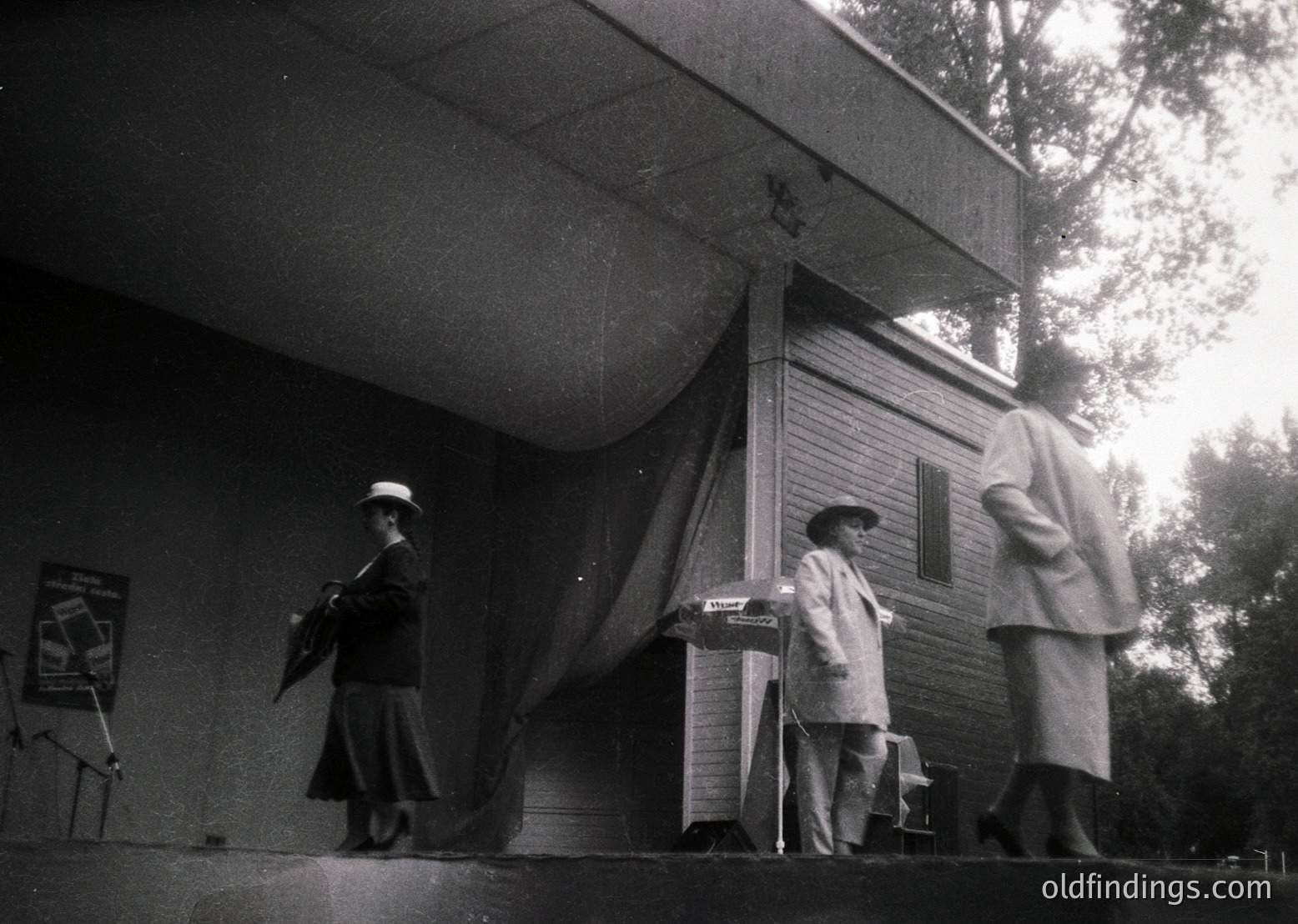 Mid-century outdoor stage performance with three musicians in 1950s-60s attire. Woman on left plays guitar; seated man at center conducts or sings with sheet music; standing man on right wears a coat and holds a mic stand. Wooden stage with overhang and trees in background.