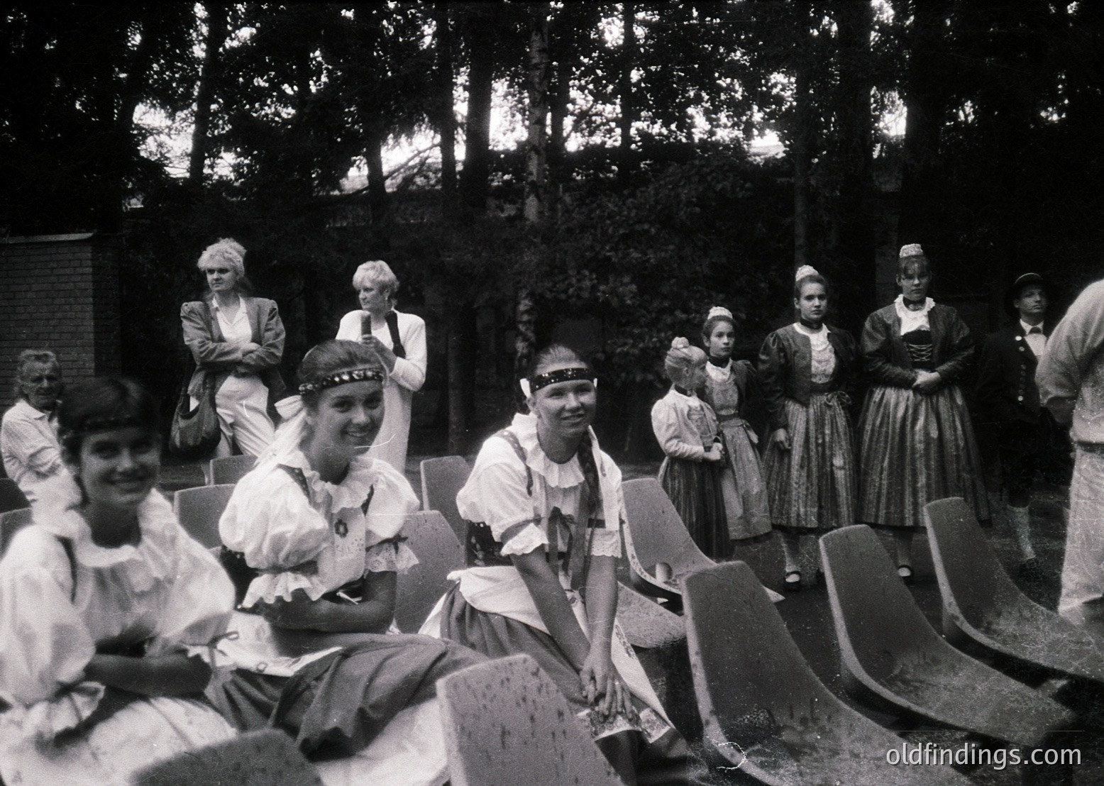 Vintage black-and-white group photo featuring women in traditional folk attire, likely Eastern European, with embroidered blouses, aprons, and headscarves. Seated and standing in an outdoor setting with wooden bleachers and dense forest backdrop. Mid-20th century (1940s–1960s) cultural or folk festival scene.