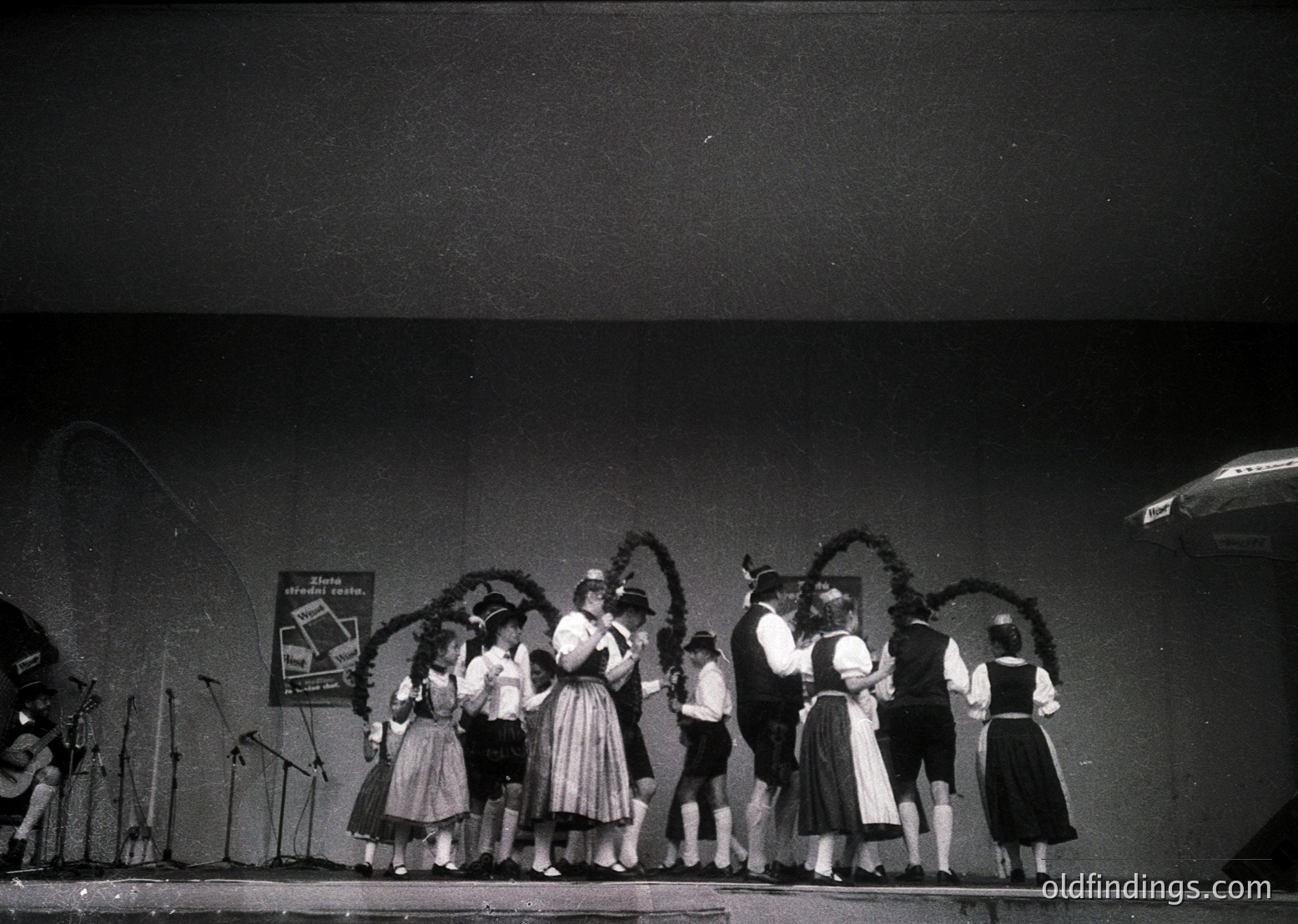 Black-and-white stage performance featuring traditional Alpine folk attire—women in full skirts, blouses, and headscarves, men in vests, knee-length pants, and hats. Stage backdrop includes floral garlands and a banner with Cyrillic text. Microphones and a microphone stand suggest live vocal performance. Likely Eastern European folk music scene, 1960s–1980s.