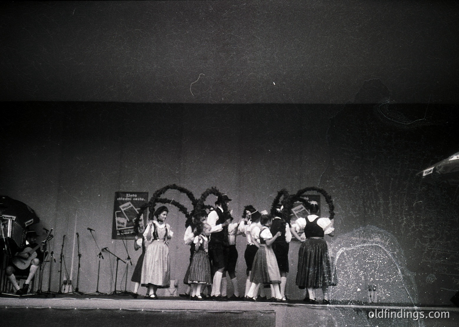 Five women in traditional Bavarian attire perform on stage, holding decorative wreaths. Stage lighting and microphones suggest a live cultural event. Likely 1950s–1970s, Germany/Austria.