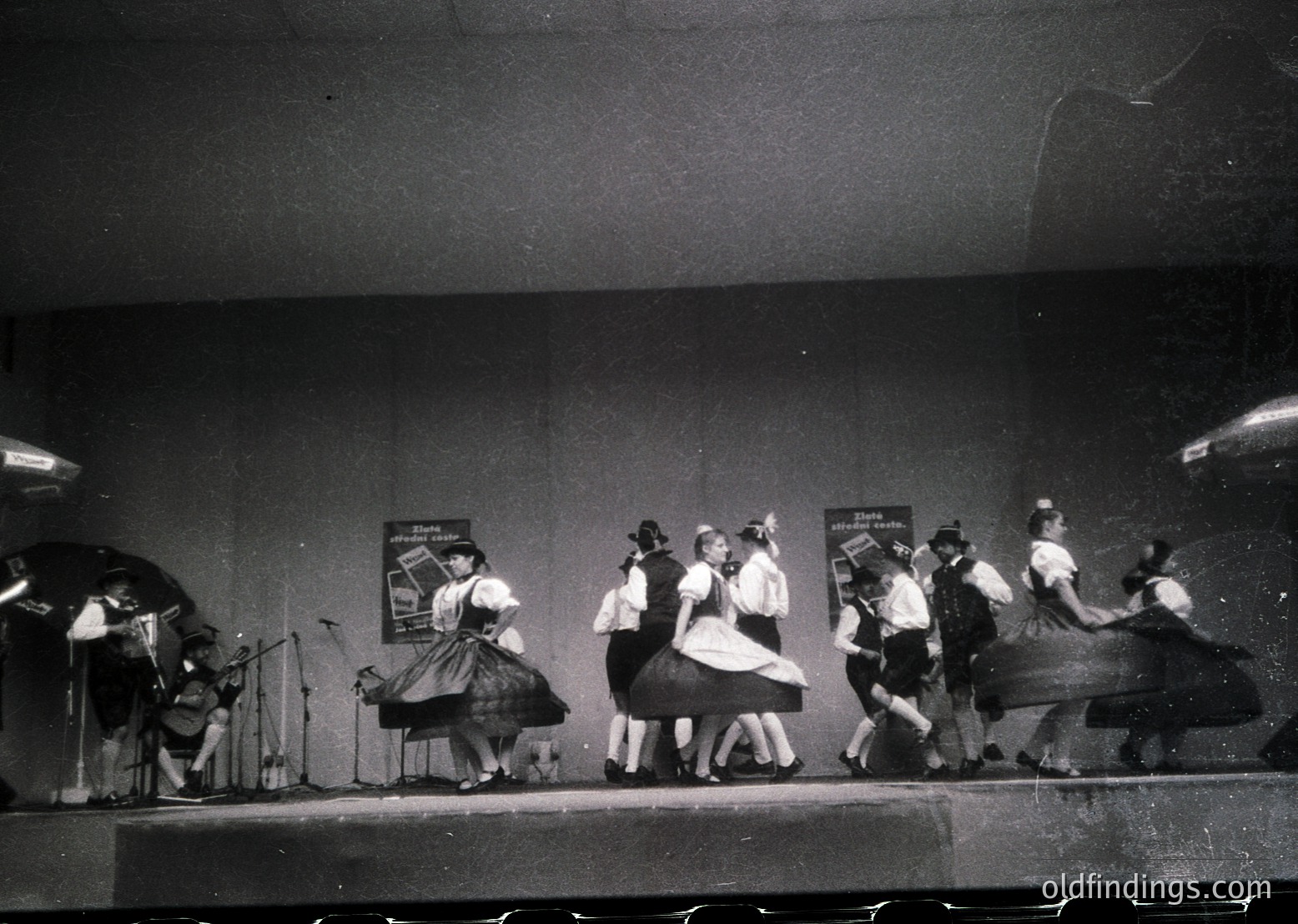 Group of dancers in traditional folk attire performs on stage, 1960s-70s. Men in vests and women in full skirts, accompanied by a musician playing accordion. Stage backdrop features framed posters.