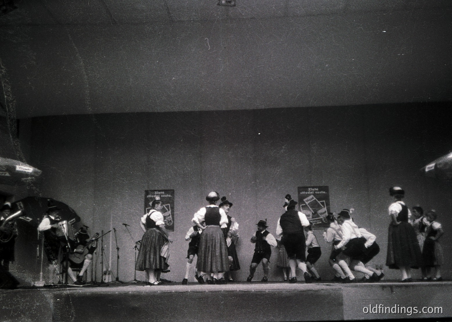 Mid-20th century stage performance featuring traditional folk dancers in regional attire, likely Eastern European. Group includes musicians (accordion, violin) and dancers in coordinated skirts, vests, and headscarves. Stage backdrop shows framed posters, suggesting a cultural or folk festival setting.