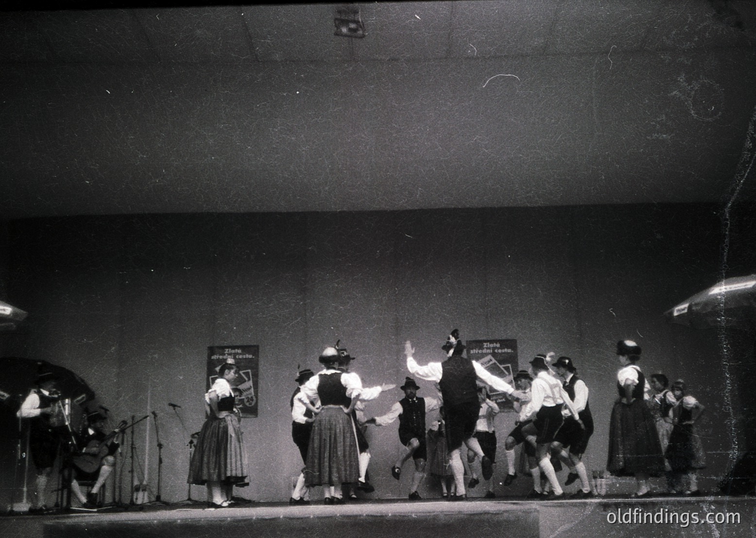 Group performing traditional folk dance on stage, 1960s-70s. Costumes feature embroidered aprons, wide skirts, and headscarves. Stage backdrop includes a banner with Cyrillic text ( )