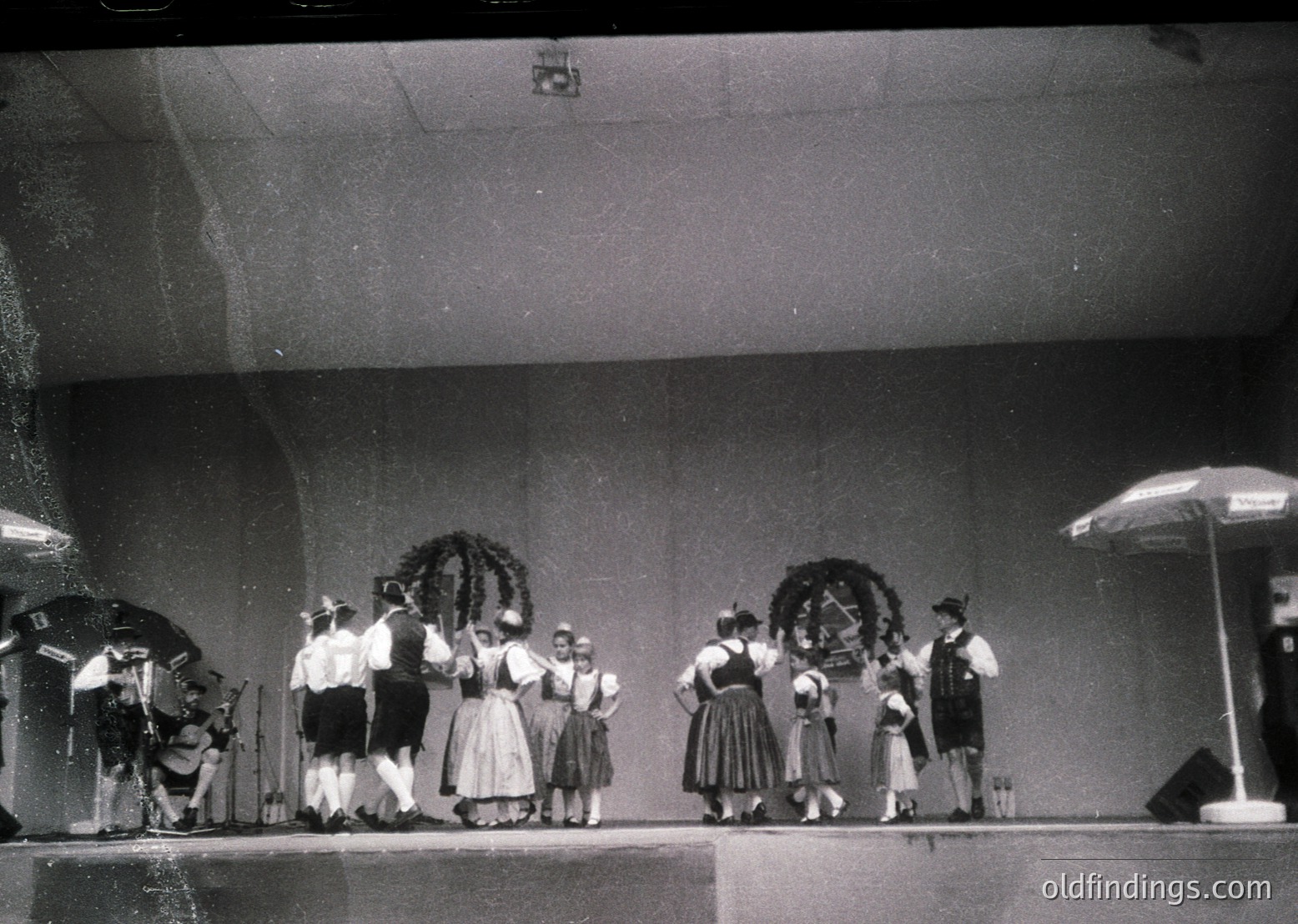 Group in traditional Alpine attire performing on stage, likely 1950s–1970s. Men wear vests, hats, and aprons; women in dirndls with floral headpieces. Stage lighting and umbrellas suggest an indoor event.
