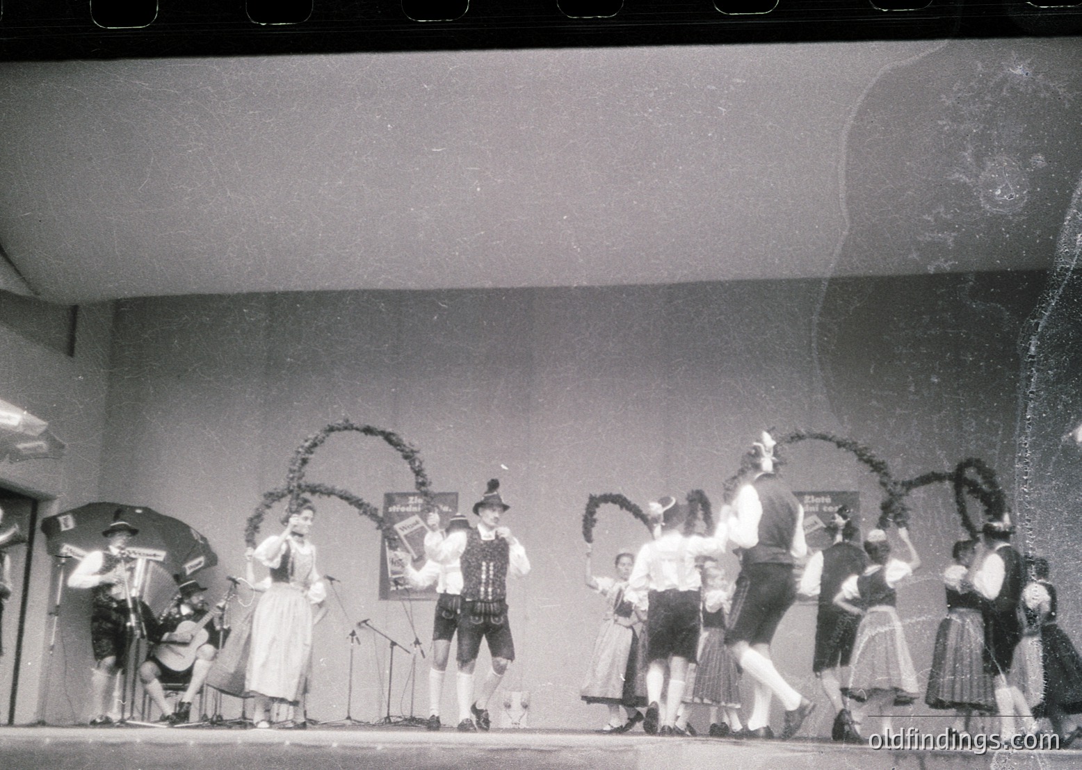 Mid-20th century folk dance performance in traditional Alpine attire. Stage setup includes floral arches, a microphone stand, and a banner with text ("*Bayerische..."). Dancers in dirndls, lederhosen, and hats perform in a hall with visible doors and a ceiling light fixture.