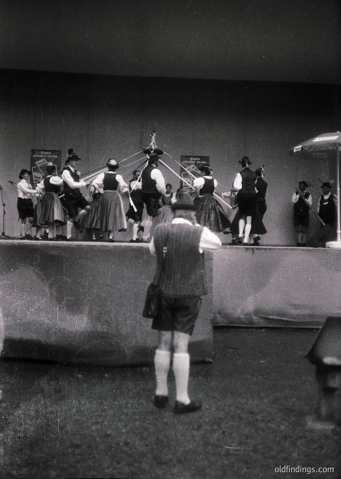 Indoor folk dance performance featuring traditional attire (dirndls, vests, hats) and musicians playing accordions. Stage setup includes microphones and a central decorative pole. Likely 1950s–1970s European cultural event.
