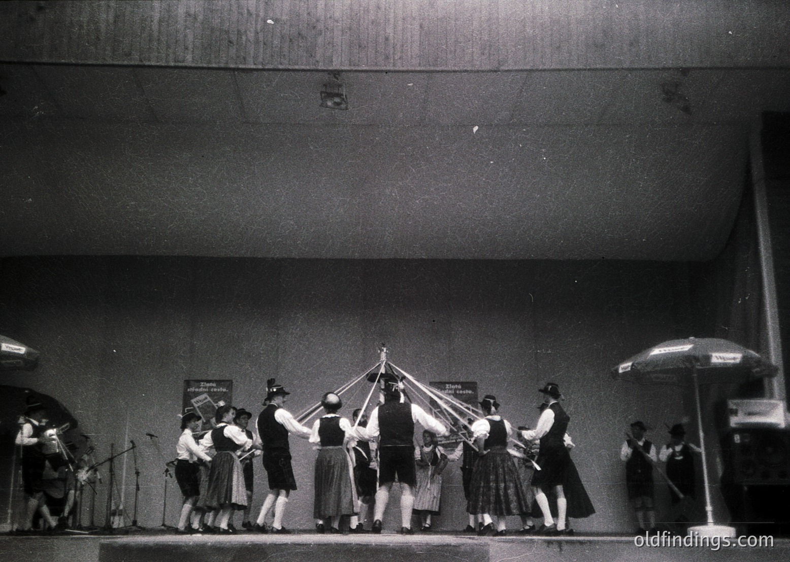 Vintage black-and-white photo of a traditional folk dance performance, likely Eastern European. Group of dancers in coordinated skirts and blouses, holding hands in a circle around a central pole. Musicians with accordions and drums positioned to the left. Stage setup includes a large umbrella and visible lighting rig.