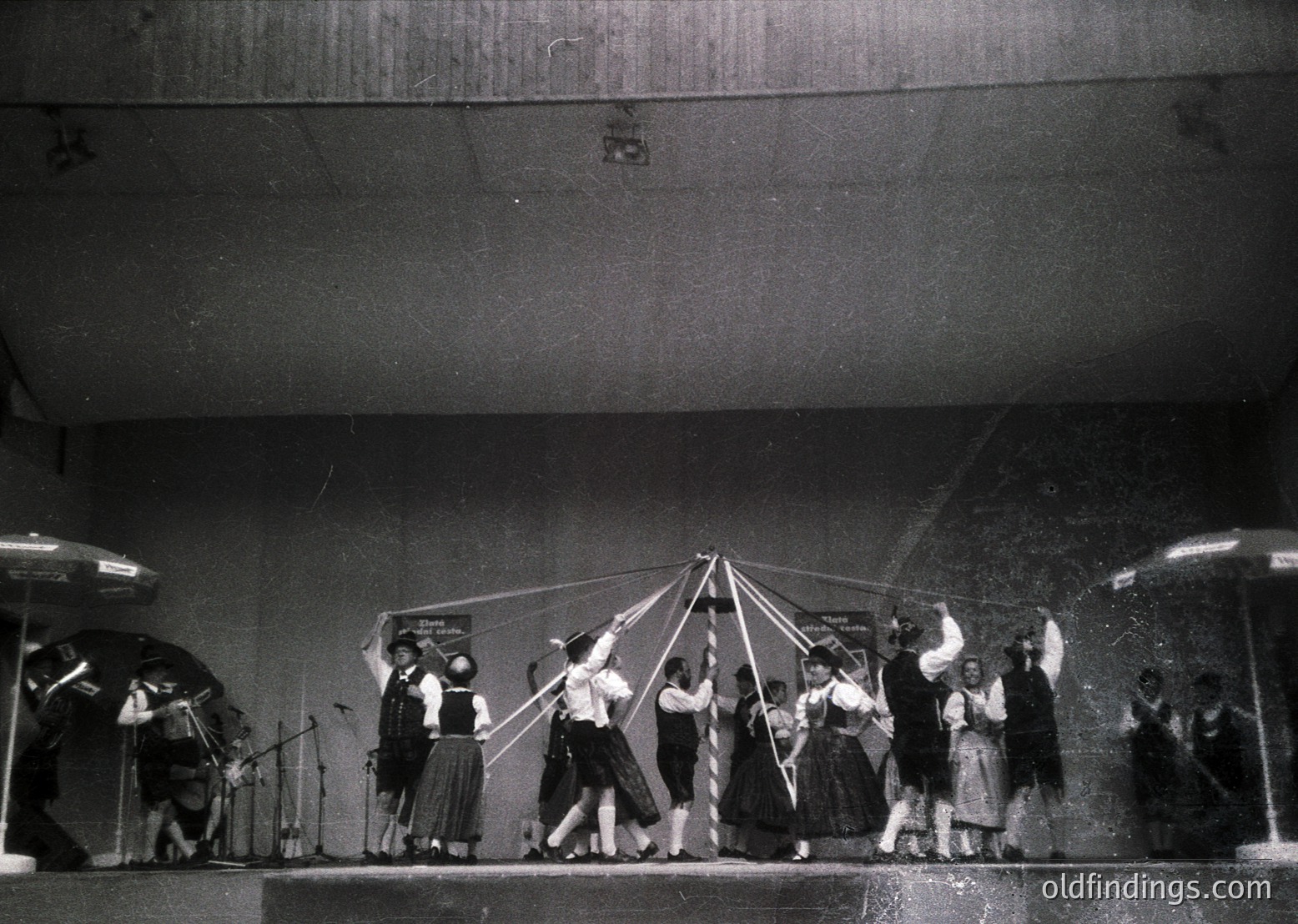 Black-and-white stage performance featuring a group of dancers in coordinated 1950s-60s attire, holding a large triangular structure. Stage lighting highlights dynamic poses; musicians play in background. Likely a folk or theatrical dance performance.
