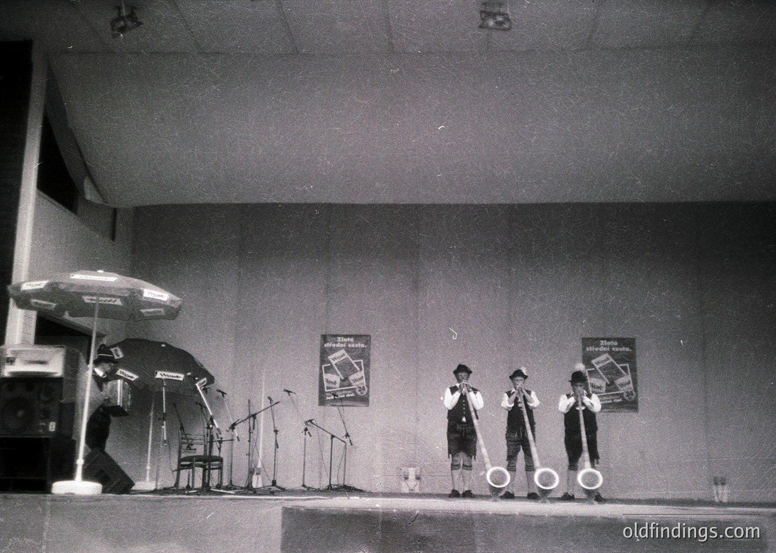 Four male performers in matching suits and hats play instruments on a stage with microphones, microfiche posters, and a drum set. Industrial lighting and a black umbrella add to the vintage concert atmosphere. Likely 1960s–1970s, Eastern Bloc or Soviet-era venue.