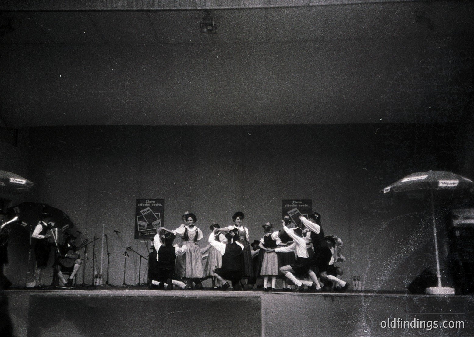 Mid-20th century stage performance featuring a group dance with musicians playing brass instruments. Women in knee-length dresses and men in suits perform choreographed steps under stage lights. Signs in Cyrillic suggest Eastern European setting (, , , , )