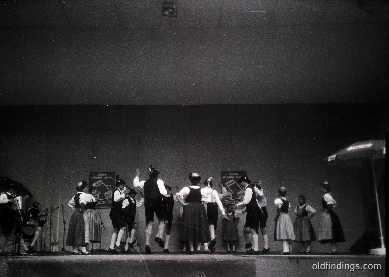 Black-and-white stage performance featuring traditional folk dancers in coordinated attire—skirts, blouses, and headscarves—with handwritten signs reading *"Danza tradicional"* and *"Danza española."* Stage lighting and a spotlight highlight the performers. Likely mid-20th century (1950s–1960s) cultural event or festival.