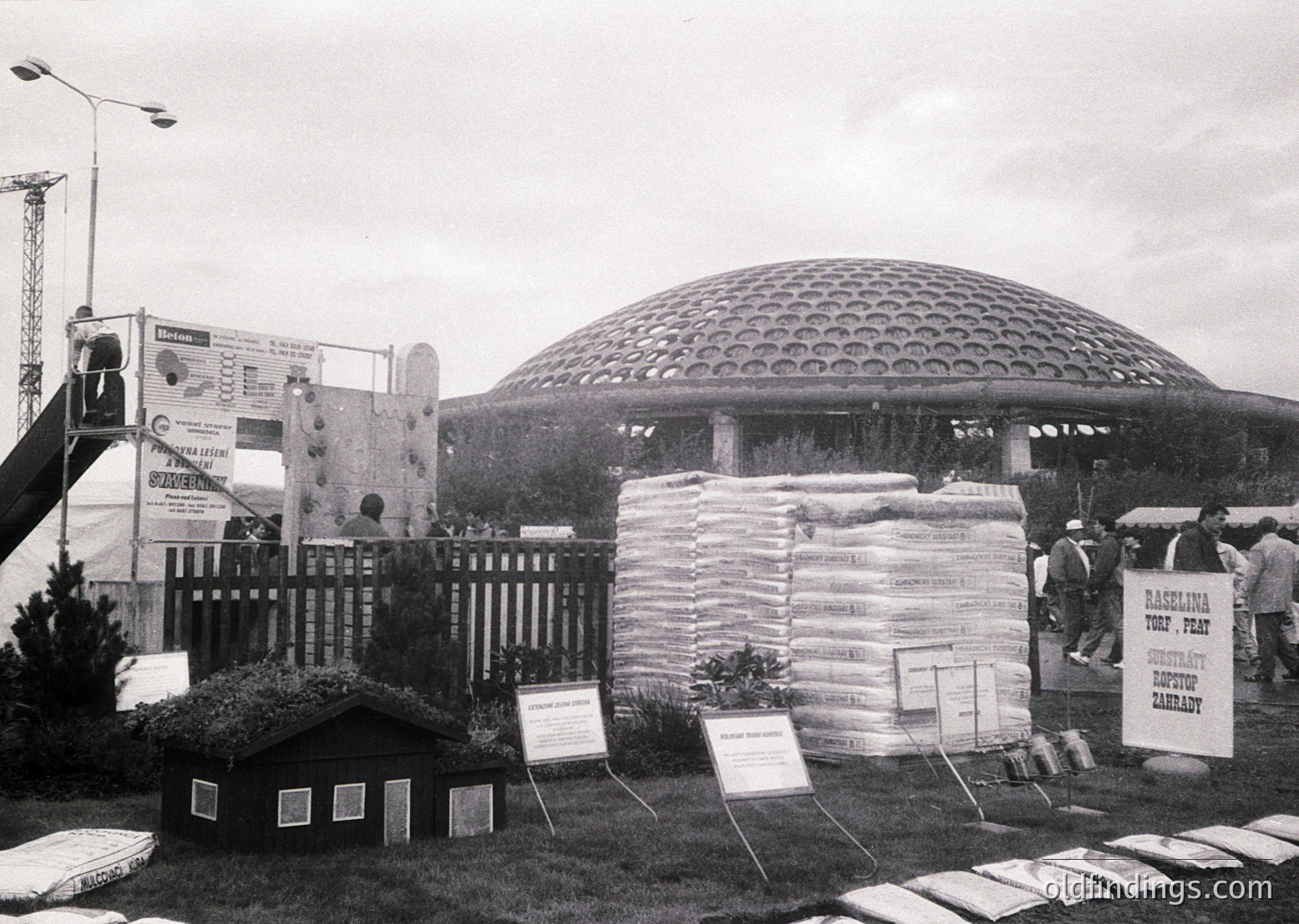 Mid-century futuristic pavilion with honeycomb dome structure, likely from a 1960s–1970s world’s fair or expo. Surrounding displays feature stacked burlap sacks labeled with agricultural products (e.g., tobacco, flax). Construction crane and promotional banners suggest industrial or agricultural focus. Crowd of visitors interacts with exhibits.