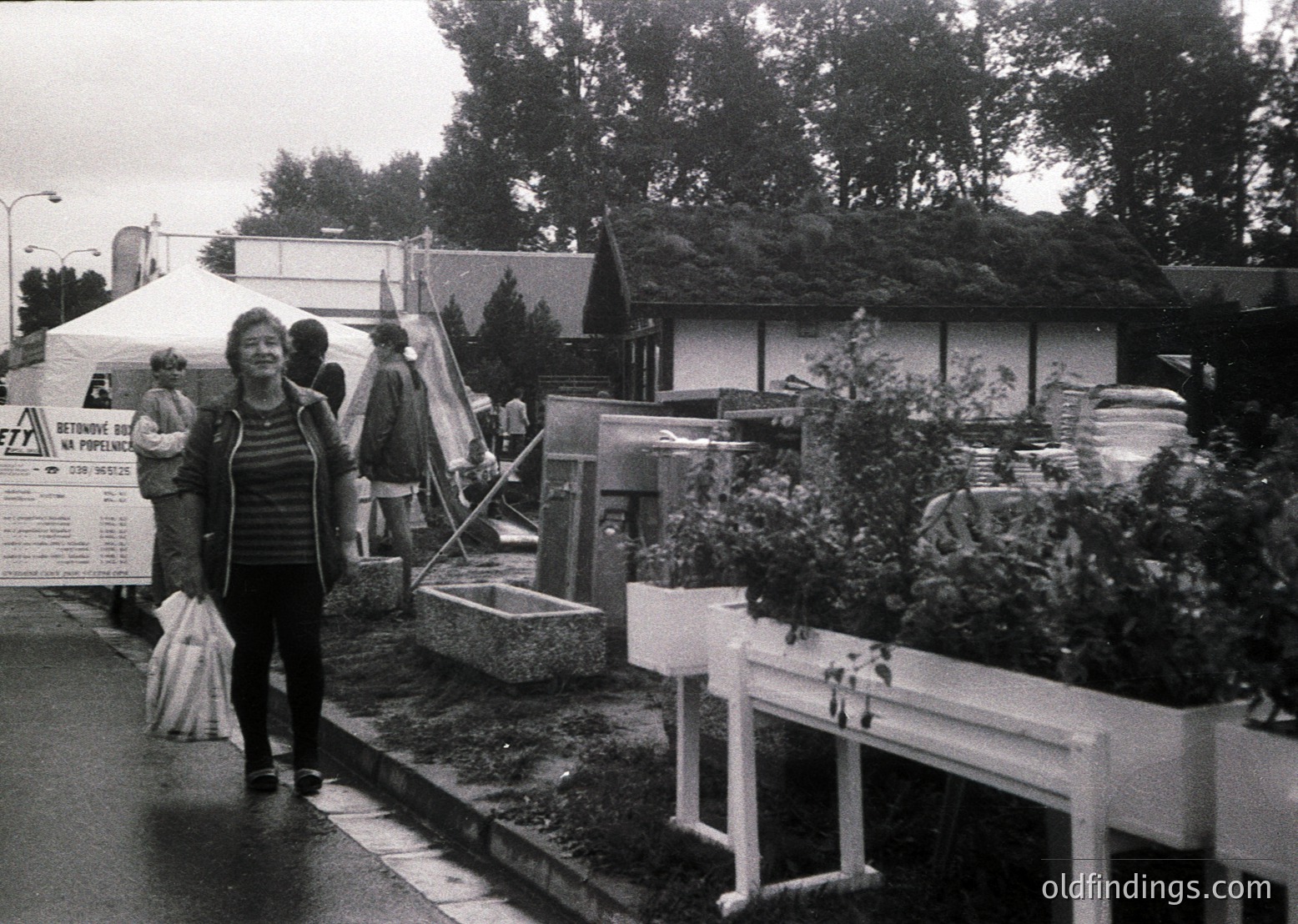 Black-and-white street scene featuring a woman in a striped sweater carrying bags, walking past a temporary market stall with a "BETON" sign. White planters with greenery line a curb, while tents and a small structure with a sloped roof occupy the background. Likely a 1970s European outdoor market or fair.