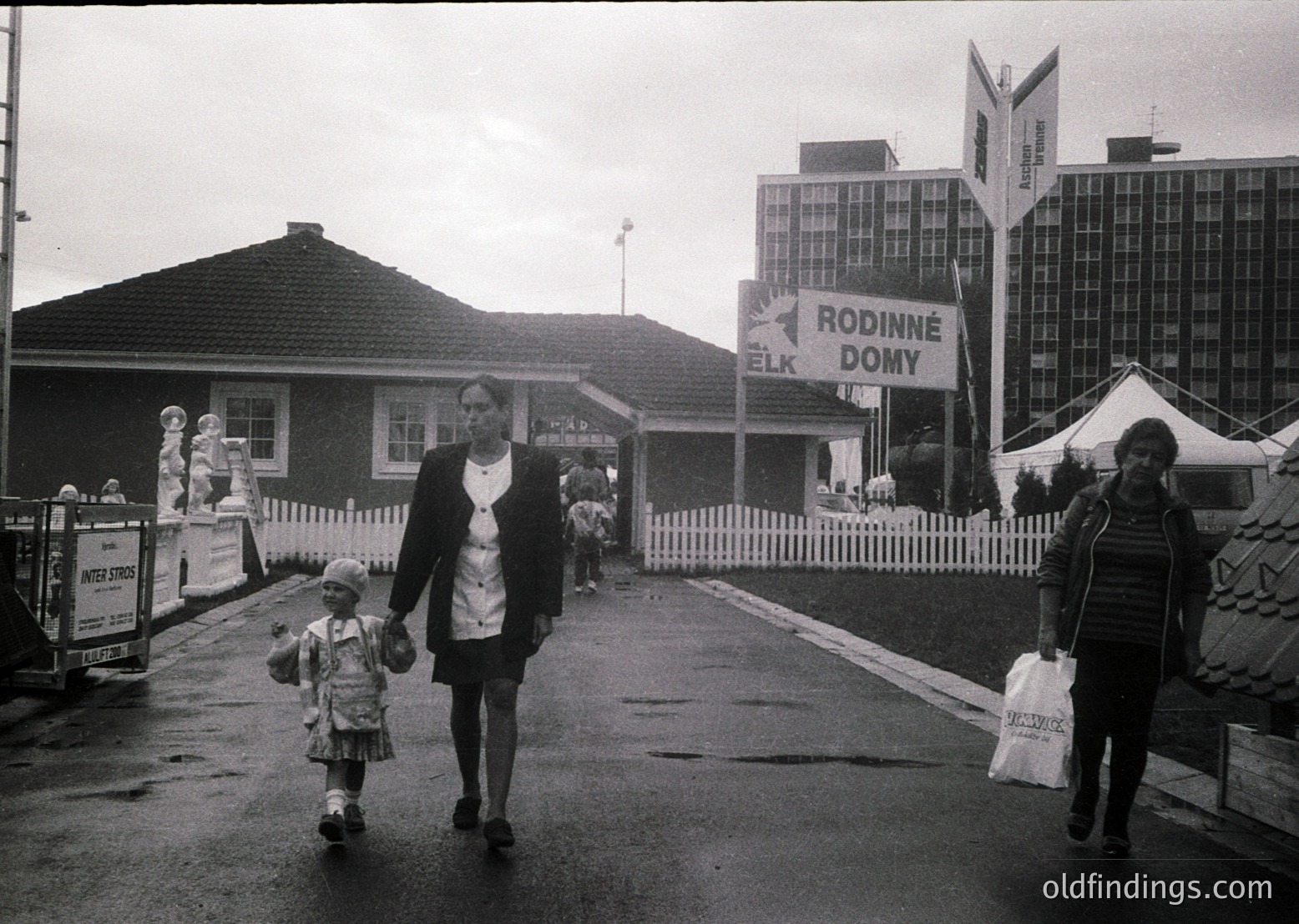 Black-and-white street scene featuring a woman in a striped coat and hat holding a child’s hand, walking toward a "Rodinné Domy ELK" housing complex. Mid-20th century urban architecture with Soviet-era concrete apartment blocks and a smaller brick building in foreground. Wet pavement suggests recent rain.