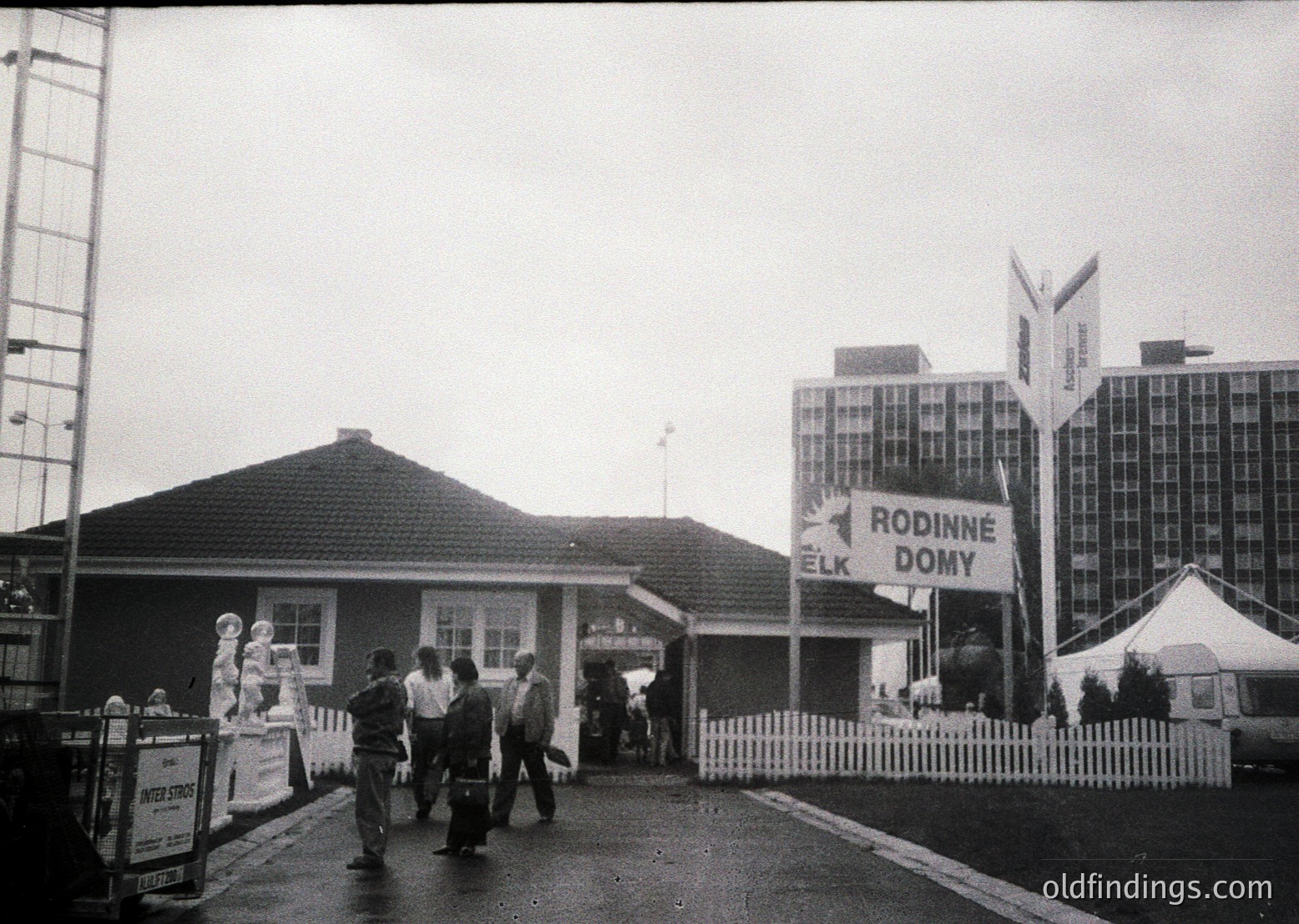 Black-and-white shot of a 1960s-era Czech family resort entrance, featuring a small wooden building with a peaked roof and a larger modernist hotel complex in background. Prominent signage reads "Rodinné Domy ELK," indicating family holiday homes. Crowd of casually dressed travelers with luggage suggests a travel or tourism setting.