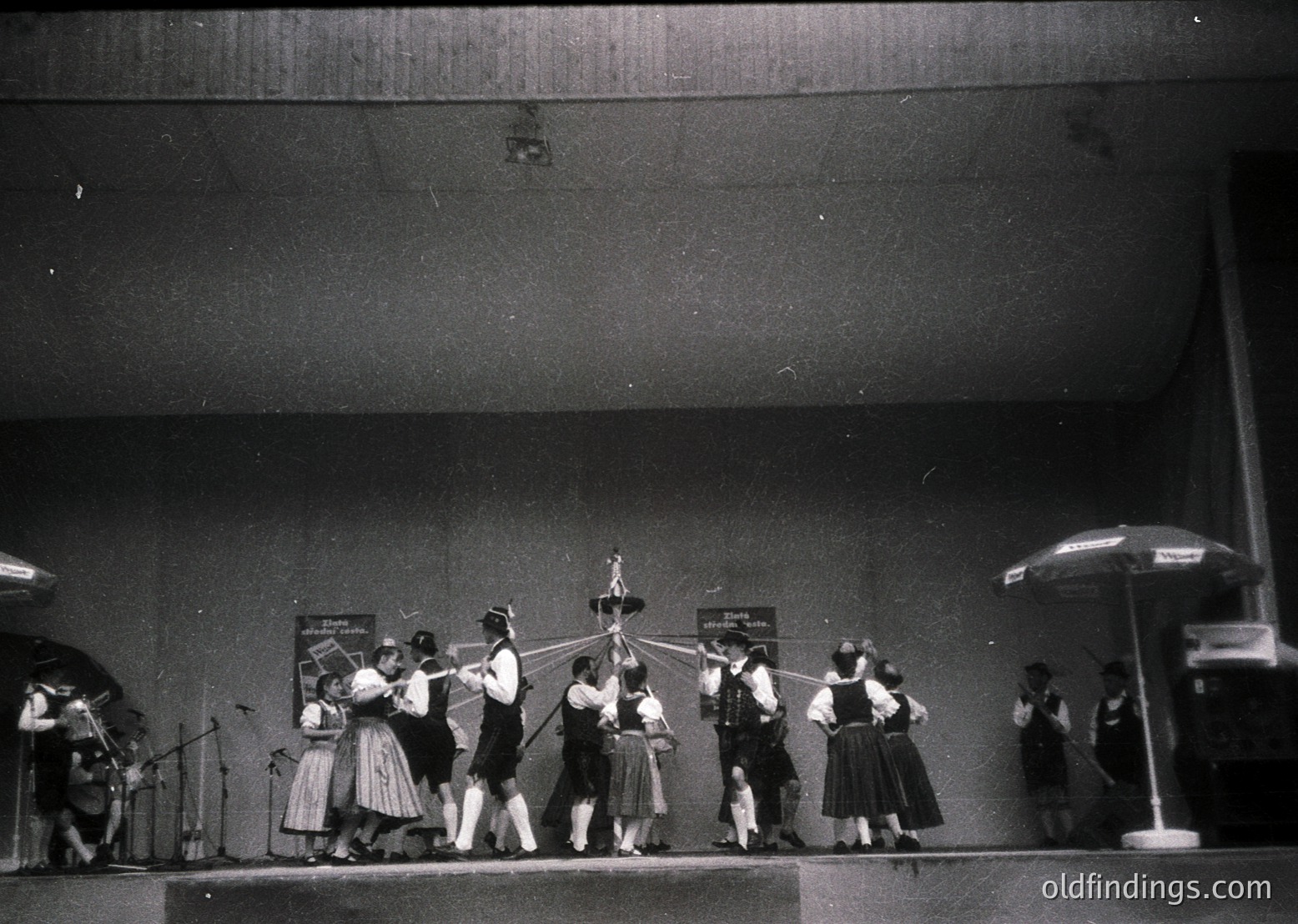A black-and-white stage performance featuring a group of dancers in traditional folk attire, likely Eastern European, performing a line formation. Stage lighting highlights the performers against a dark backdrop with visible posters. Microphones and a small band section (violin, accordion) flank the dancers. Umbrella props suggest a rain-themed or symbolic motif. Mid-20th century (1950s–1960s) cultural or folk festival setting.