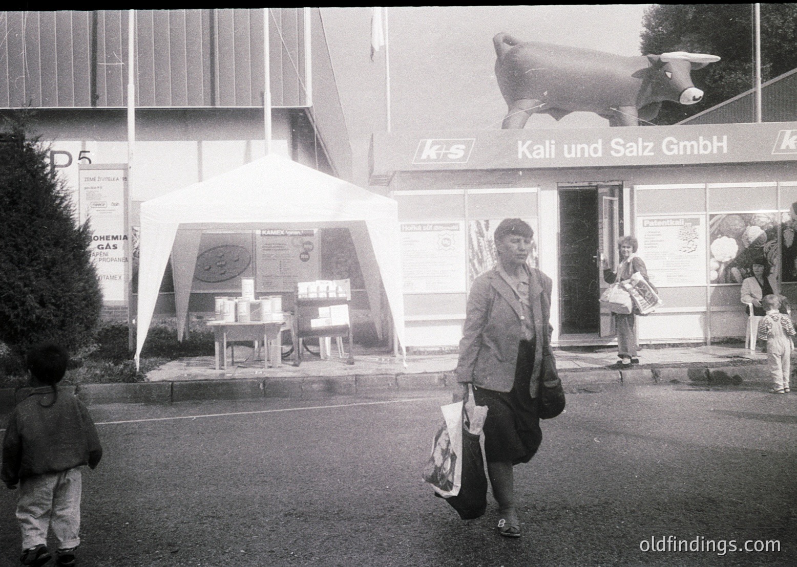 Mid-20th century street scene outside a **Kali und Salz GmbH** storefront, likely Germany. A woman in a structured suit carries shopping bags; a child stands nearby. Prominent cow logo and signage reflect 1960s-70s commercial design. Industrial architecture with geometric signage.
