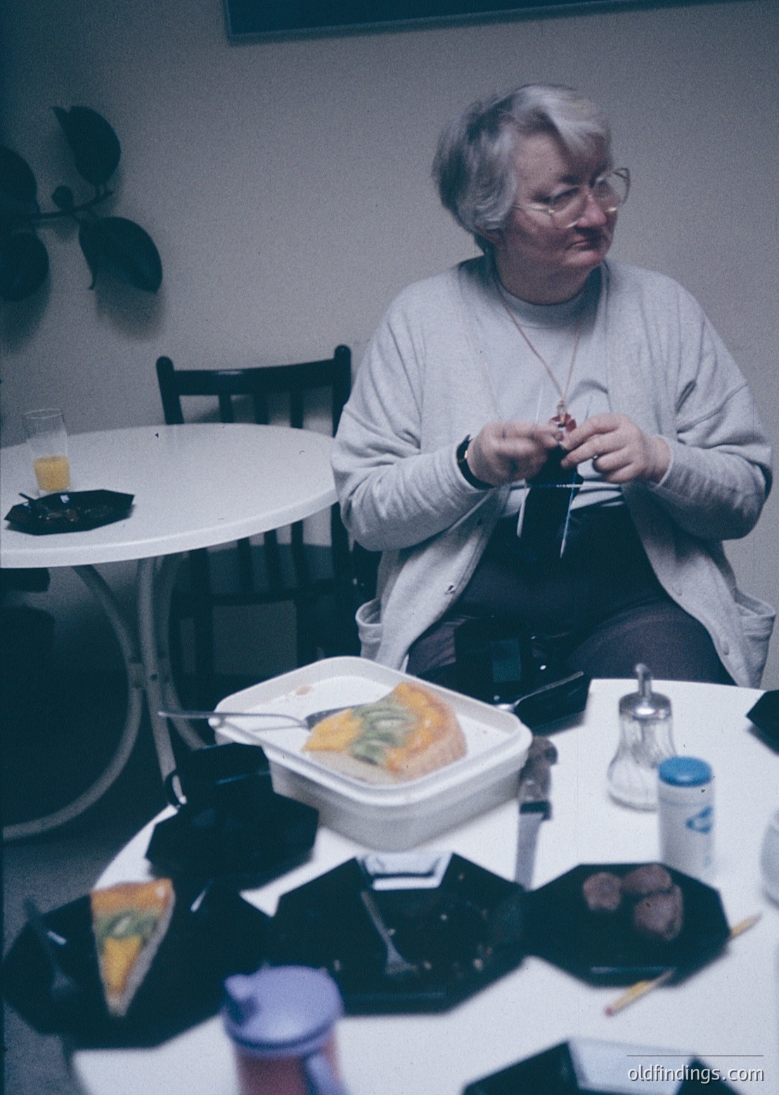 Mid-century indoor dining scene featuring a woman in a light sweater, seated at a round table with a white tablecloth. She holds a fork and knife, preparing a meal from a takeaway container. Surrounding items include a plastic container of food, a glass of water, a salt shaker, and a small bottle of condiment. The setting suggests a casual, home-cooked meal in a retro-style kitchen or dining area.