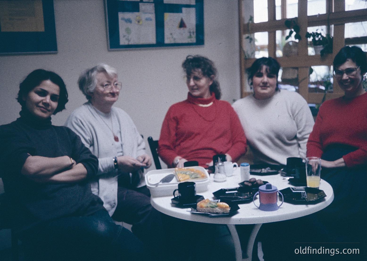 Four women seated around a round table in an indoor café, likely from the **1980s–1990s**. Warm lighting, retro decor with framed posters, and casual attire (sweaters, glasses) suggest a relaxed social gathering. Tabletop includes disposable cups, a muffin, and a coffee pot—common in mid-century dining culture. éCulture