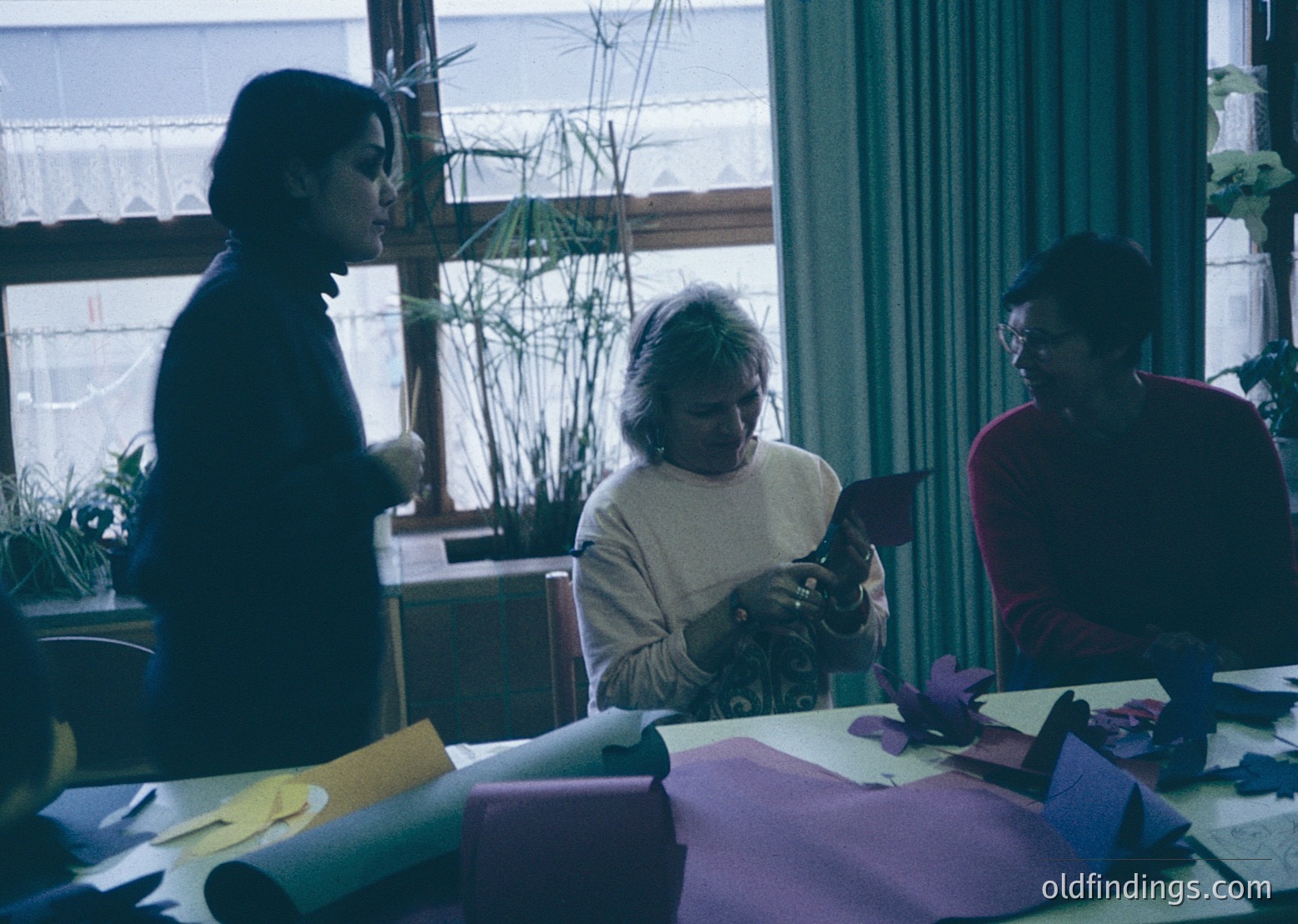 Three individuals collaborate on a craft project in a bright, indoor setting, likely a 1970s–1980s classroom or community center. Natural light floods through large windows with wooden frames, illuminating rolled fabric, scissors, and paper cutouts on a table. The woman on the left wears a dark sweater, the center figure a light sweater, and the right a red top. Decorative plants frame the window, adding warmth.