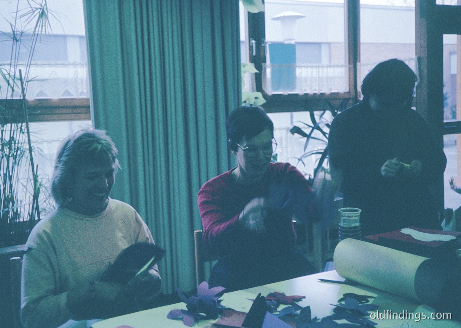 Vintage group crafting session indoors, likely 1970s–1980s. Three women seated at a table cutting paper shapes, surrounded by craft supplies. Soft lighting, floral-patterned curtains, and a potted plant add warmth. Urban backdrop visible through windows.