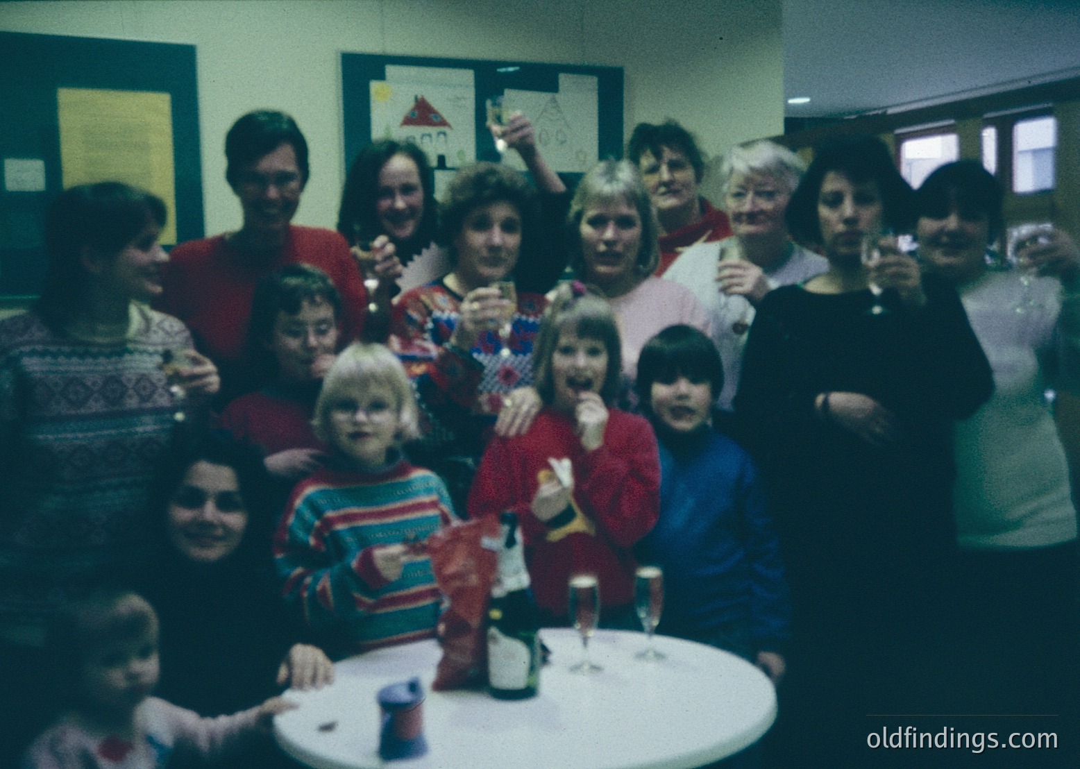 Vintage group photo from the 1980s featuring adults and children in festive attire, likely a holiday gathering. Indoor setting with a table holding champagne flutes and a small Christmas tree. Warm, retro lighting and 1980s fashion (striped sweaters, cardigans) visible.