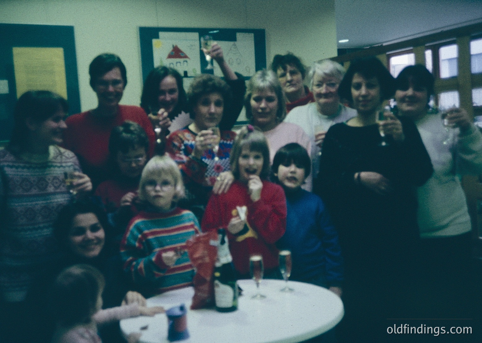 Vintage group photo from the 1970s–80s, likely a community or family gathering. Indoor setting with casual attire—striped sweaters, cardigans, and festive holiday decorations. Adults and children pose around a table with wine glasses and a bottle. Warm, nostalgic lighting and slightly blurred edges enhance vintage charm.