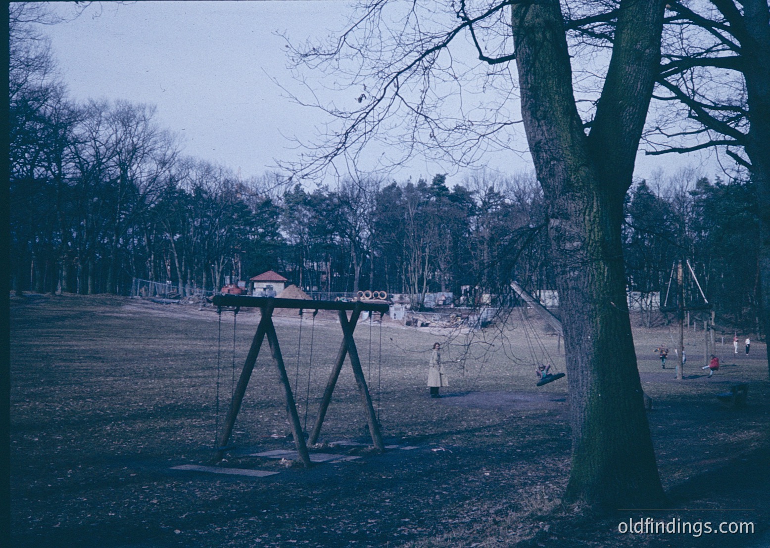 Vintage playground in a wooded park, featuring a swing set with metal frames and chains. Background shows a small pavilion and scattered trees. Likely mid-20th century (1950s–1970s) based on equipment design.