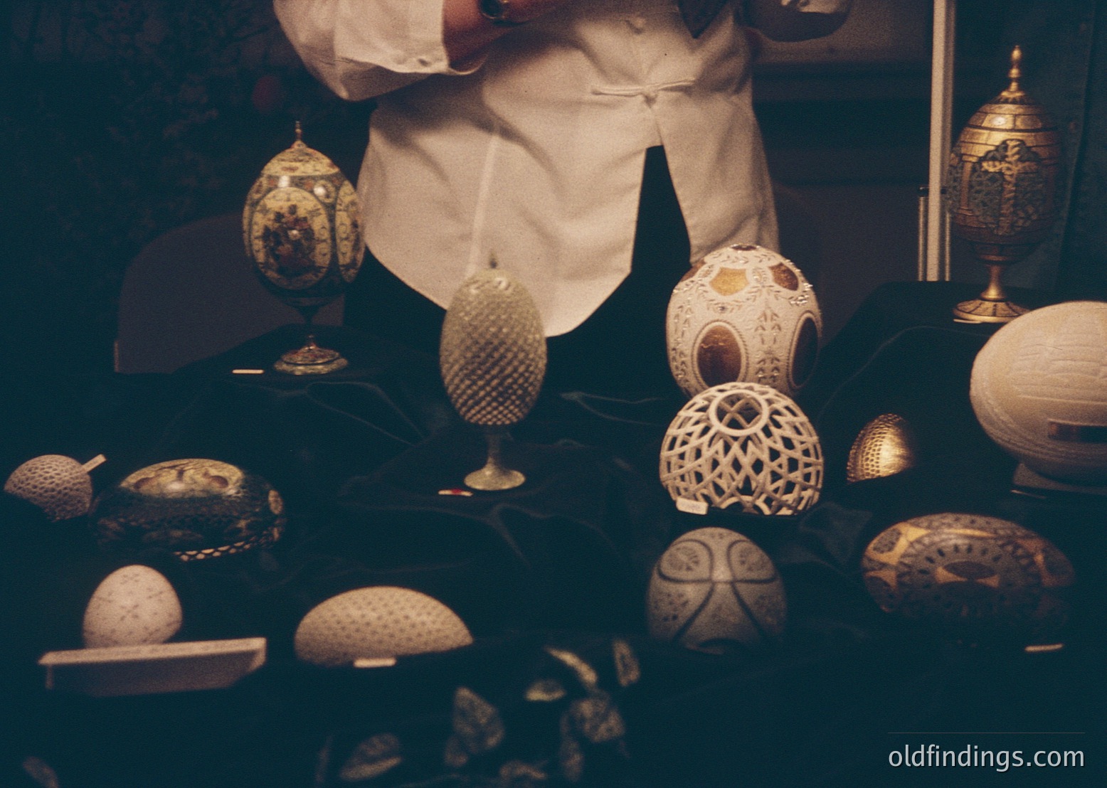 Vintage egg-shaped decorative items displayed on a dark surface, likely from a 20th-century European craft tradition. Intricate patterns include floral, geometric, and lattice designs in gold and white tones. Items appear handcrafted, possibly ceramic or porcelain, with ornate stands. Warm sepia tone enhances nostalgic aesthetic.