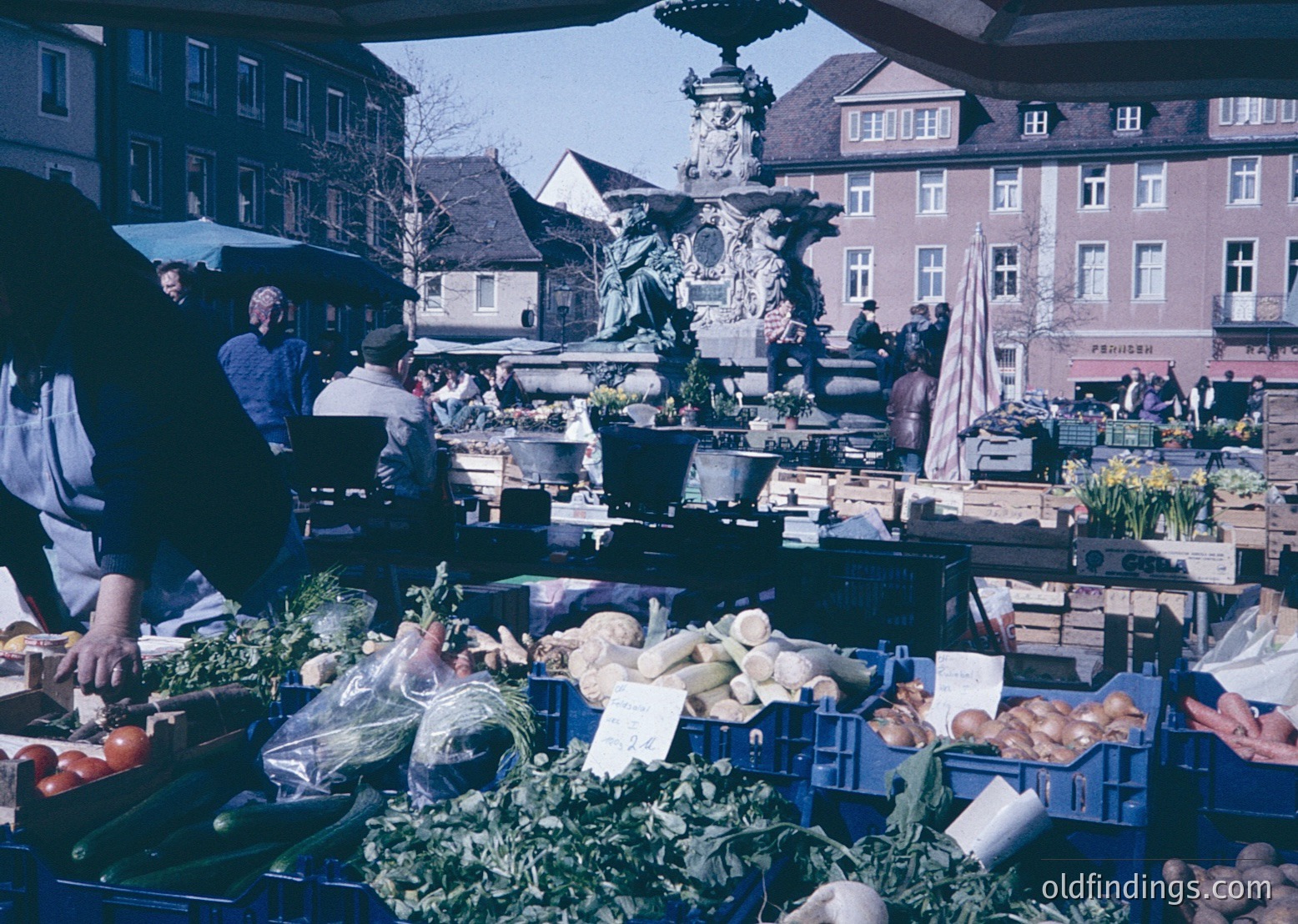 Vibrant 1970s European outdoor market with fresh produce—root vegetables, greens, and crates—displayed under blue plastic covers. Ornate fountain and historic brick buildings () frame the scene. Crowds gather around stalls, suggesting lively community engagement. (, , , , )