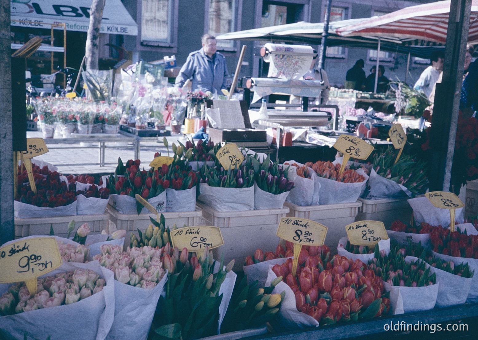 Vibrant 1960s outdoor flower market stall with tulips, roses, and carnations priced in German marks. Signs indicate "Reform" and "Kölbe" shops in background. Urban setting with pedestrians and vintage signage.