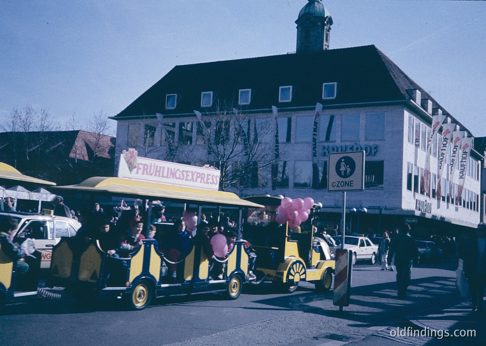 Vintage street scene featuring a yellow "Frühlingsexpress" (Spring Express) tram with passengers, likely from the 1960s–70s. The tram, adorned with pink balloons, operates in front of a white building with "Konditorei" signage, suggesting a café or bakery. Pedestrians and vintage cars populate the scene, indicating a lively urban atmosphere. é