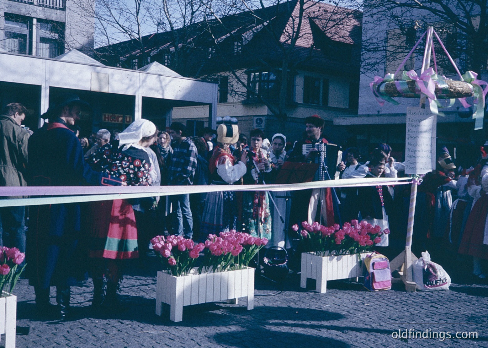 Vintage street festival featuring traditional costumes and floral displays. Crowd gathers around a stage with tulips in white planters, likely part of a spring celebration. Architectural backdrop includes a white building with a "HEART" sign. Colorful banners and a decorated pole add festive atmosphere. Likely European, 1960s-1970s era.