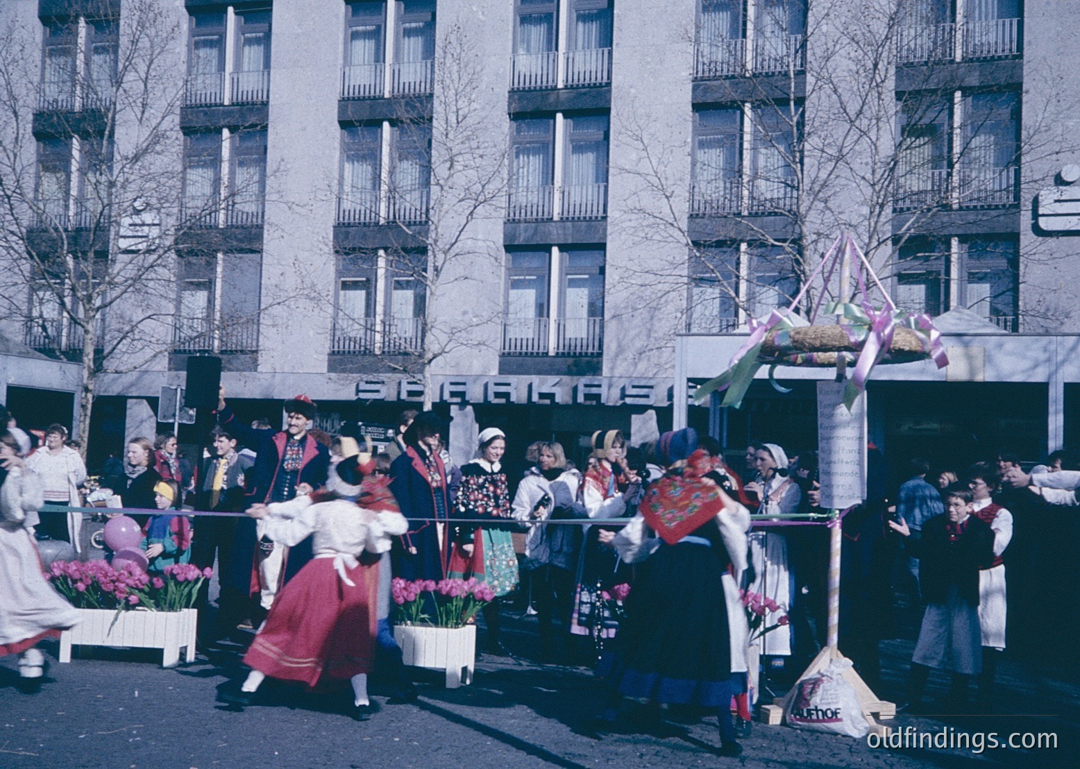 Vibrant street parade featuring traditional folk dancers in colorful, embroidered costumes, likely Eastern European. Barrels of tulips line the street, suggesting springtime celebration. Mid-20th century urban setting with Soviet-era apartment buildings. Crowd gathers along roped-off area, indicating a public festival or holiday event.