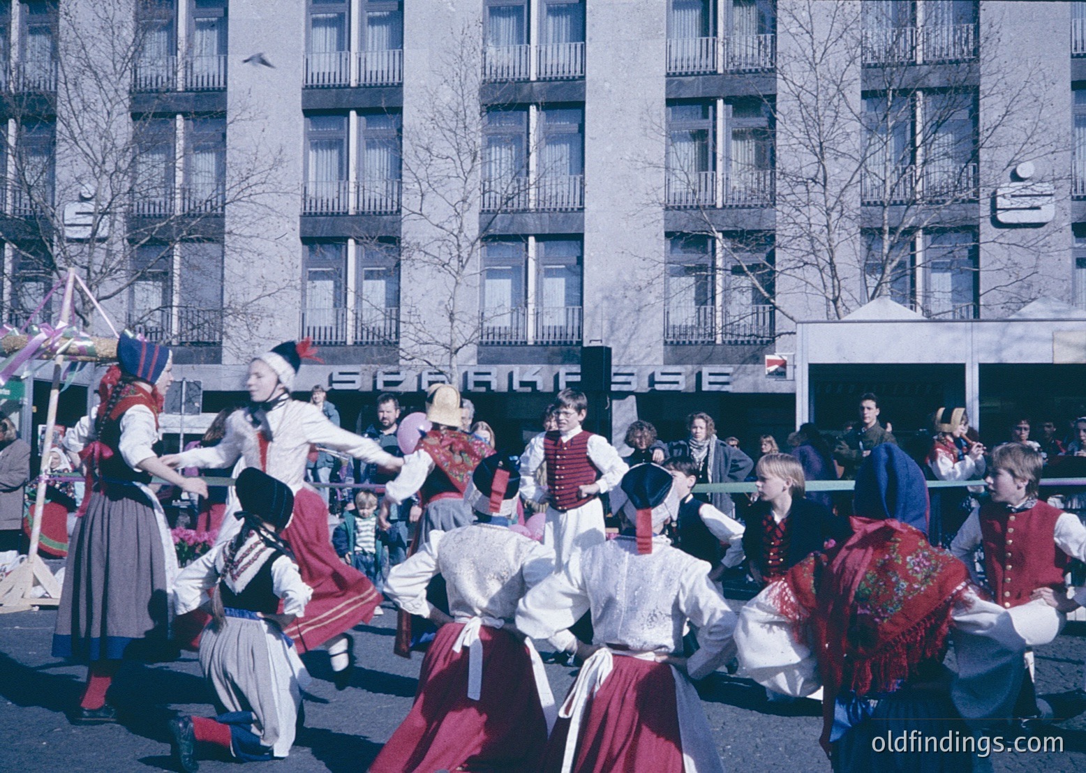 Traditional folk dancers in vibrant, embroidered costumes perform in an urban square, likely Bulgaria, 1960s-70s. Men wear red sashes and black hats; women display intricate white blouses with red aprons. Spectators line the background behind concrete buildings with balconies.