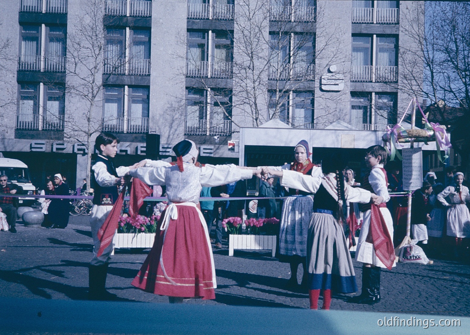 Traditional folk dancers in coordinated red skirts, white blouses, and embroidered vests perform outdoors in an urban setting, likely Eastern Europe, 1960s–70s. Multi-story concrete buildings and a crowd of onlookers frame the scene. Vibrant colors and dynamic poses capture cultural heritage and communal celebration.