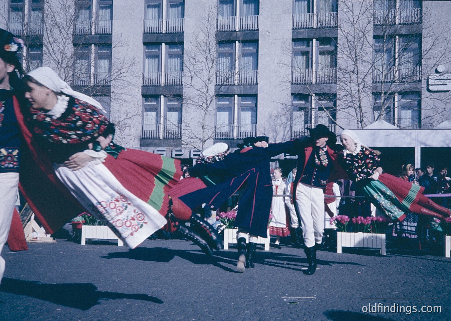 Traditional folk dancers in vibrant, embroidered costumes perform outdoors in a Soviet-era urban setting. Men in dark vests, white pants, and tall hats balance on one leg; women wear layered skirts with floral patterns and headscarves. Mid-20th century (1960s–70s) urban square, likely Eastern Bloc.