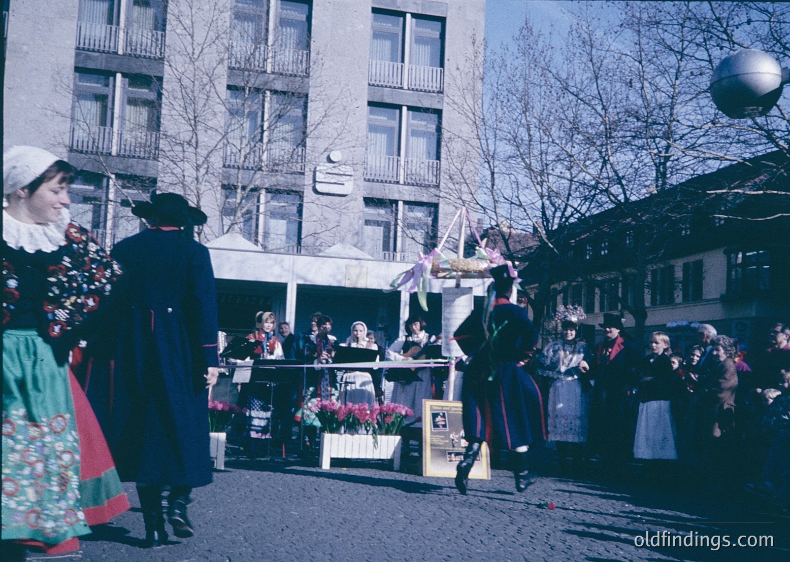 Vintage street performance featuring traditional folk dancers in colorful attire, likely Eastern European. Men in dark jackets with red trim and women in floral dresses with headscarves. Crowd gathers around a stage with floral decorations. Mid-20th century urban setting with multi-story concrete buildings.
