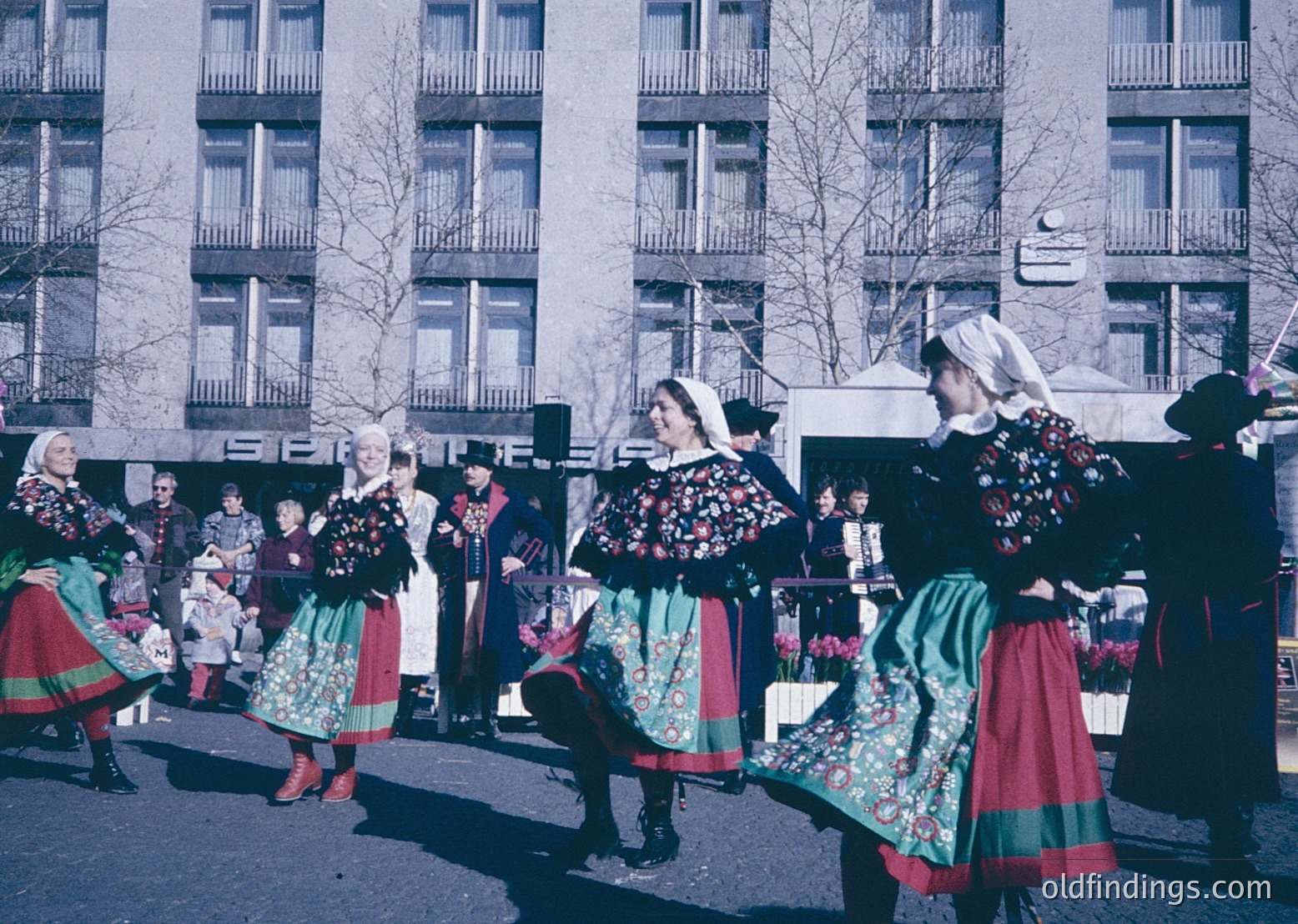 Traditional folk dancers in vibrant, embroidered costumes perform in an urban setting, likely Eastern Europe, 1960s-70s. Green, red, and floral patterns dominate their attire, paired with white blouses and headscarves. Multi-story concrete buildings and bare trees frame the scene, suggesting winter or early spring.