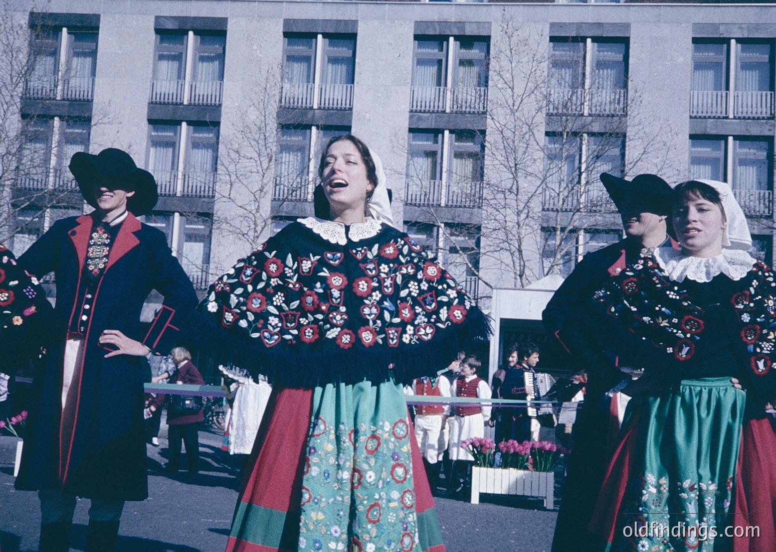 Traditional folk dancers in elaborate embroidered costumes, featuring floral patterns and layered skirts, perform outdoors. The men wear dark coats with red accents and wide-brimmed hats; women’s attire includes white collars and green skirts. Mid-20th century urban setting, likely Eastern European.