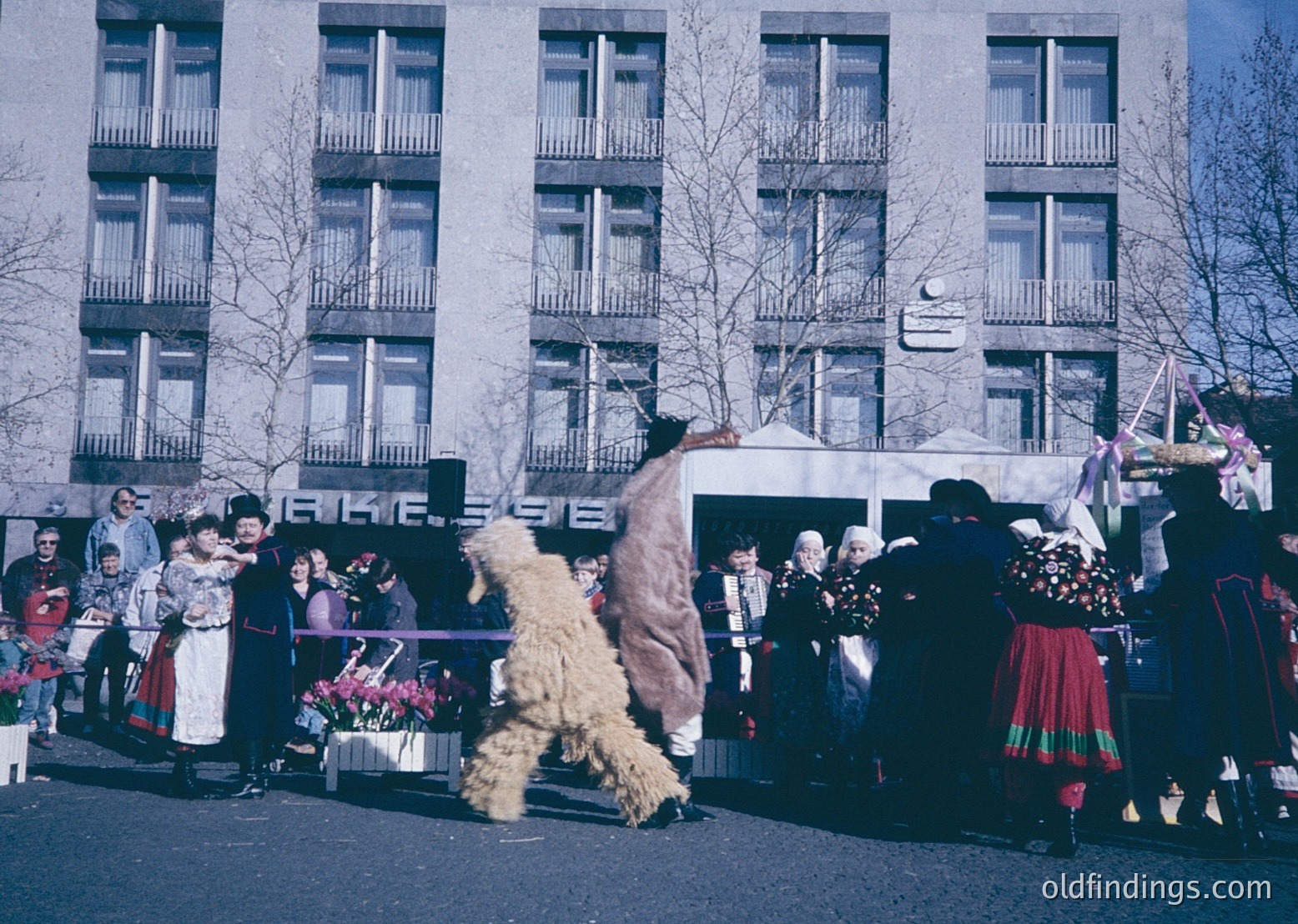 Vintage street scene featuring traditional folk performers in Eastern European attire, likely Bulgaria. A masked dancer in a shaggy costume dances near a crowd in colorful embroidered garments. Soviet-era concrete building with balconies and a crescent moon emblem in background.