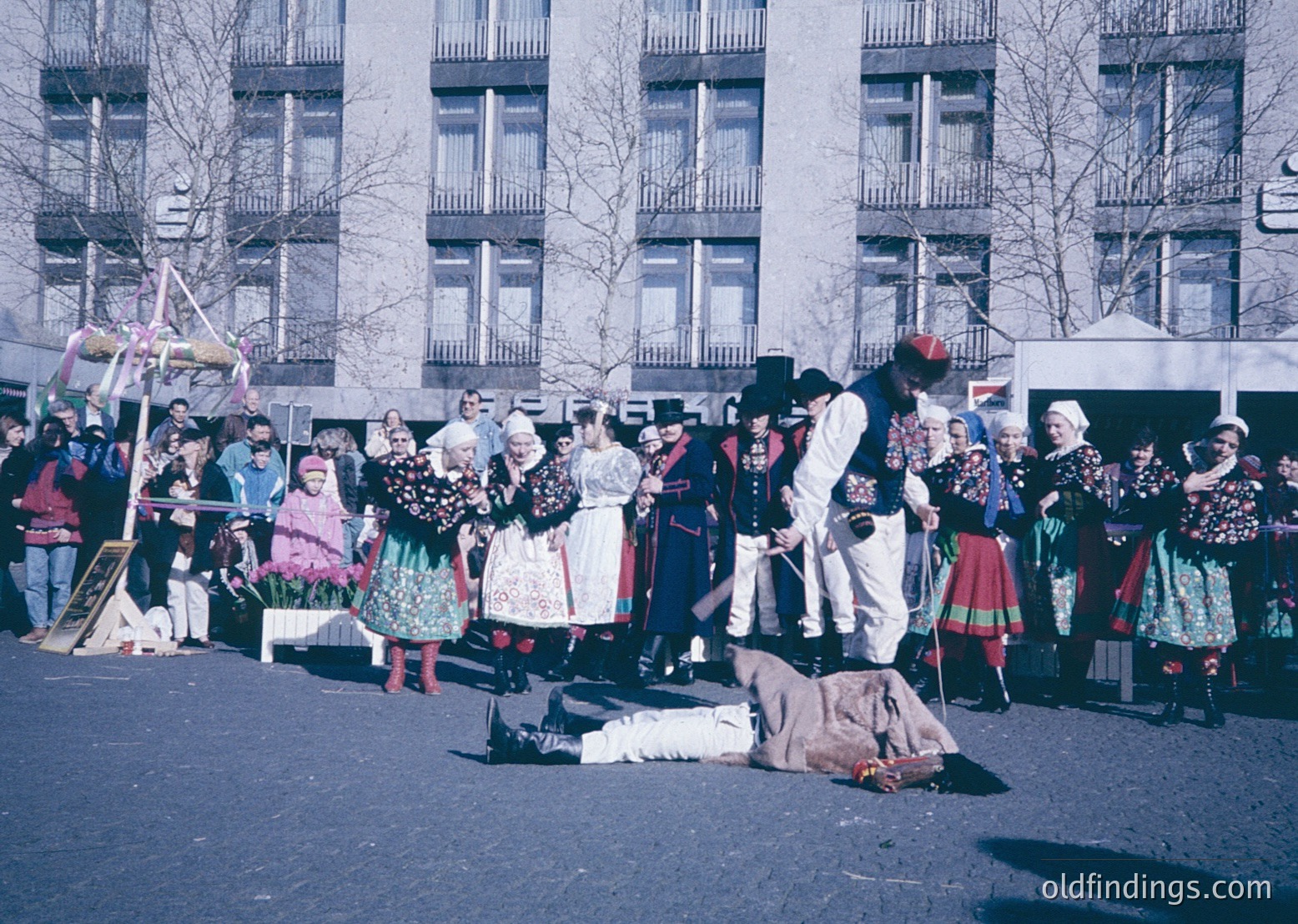 Traditional folk dance performance in urban setting, likely Bulgaria. Group in colorful embroidered costumes with men in white tunics, red hats, and women in layered skirts and blouses.