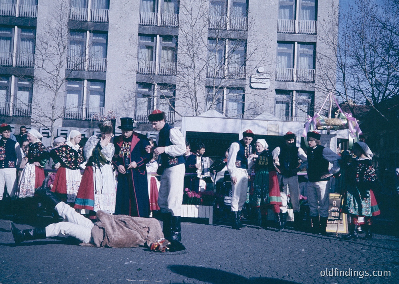 Traditional folk performance in Eastern European attire, likely Bulgaria. Group in elaborate embroidered costumes with men in vests, hats, and women in layered skirts and headscarves. Central figure in white uniform appears to lead or direct. Urban setting with Soviet-era apartment blocks in background.