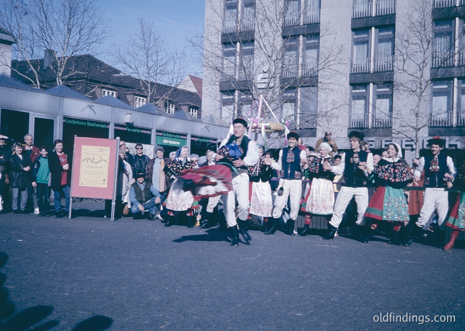 Traditional folk dancers in colorful, embroidered costumes perform in an urban square, likely during a cultural festival. The lead dancer wears a tall hat and carries a decorated pole. Mid-20th century European street scene, possibly or . Urban architecture and signage hint at a European city.