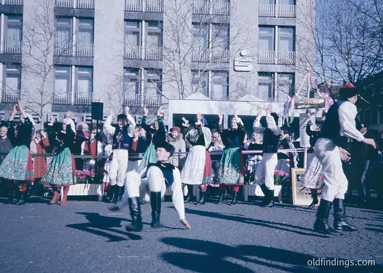 Vibrant street parade featuring traditional folk dancers in colorful, embroidered costumes—men in vests, hats, and knee-high boots, women in layered skirts and blouses—dancing in unison. Urban setting with Soviet-era concrete buildings in background. Likely 1960s–1980s Eastern Europe.
