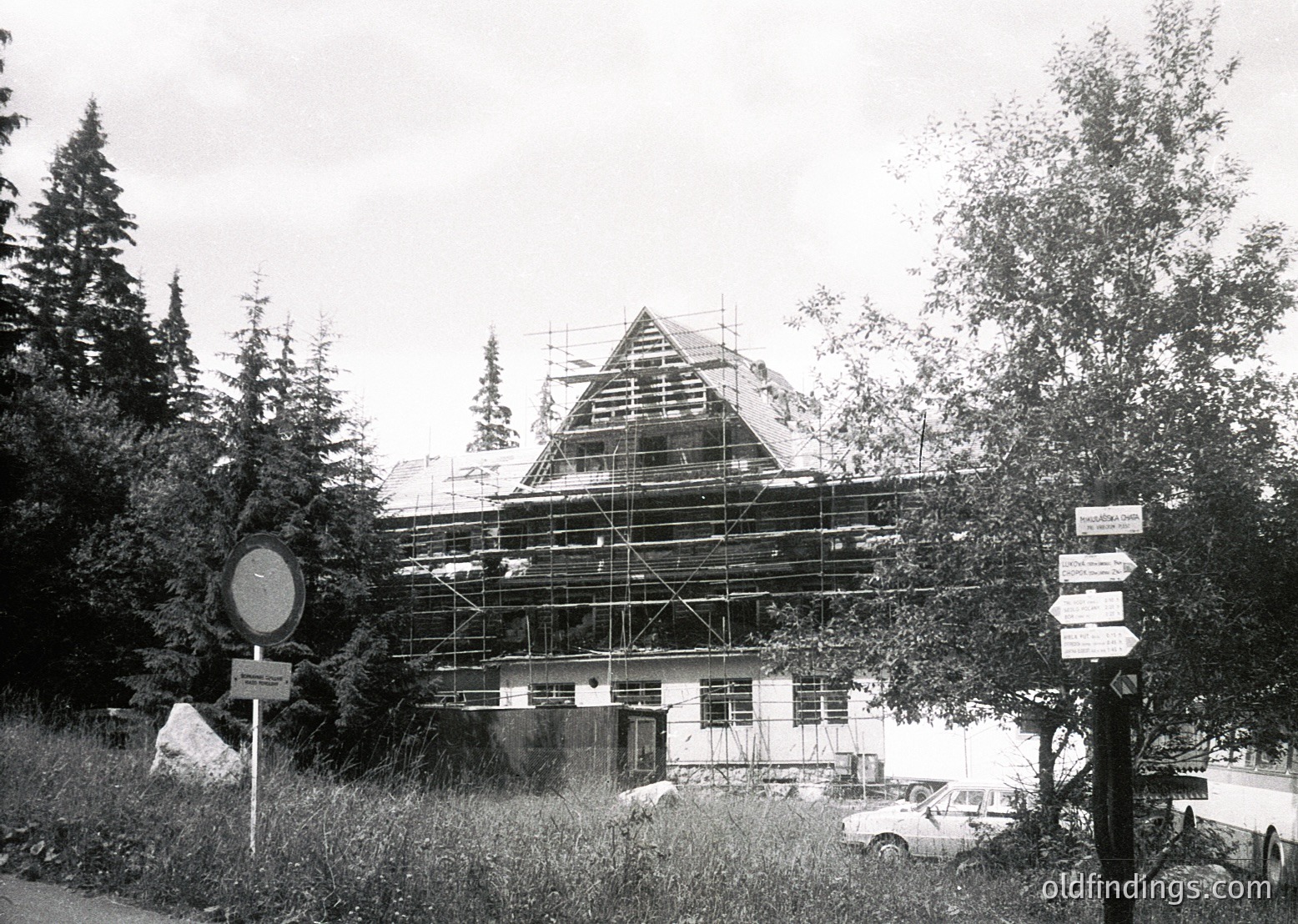 Mid-20th century alpine building under scaffolding, likely a chalet or resort. Distinctive gabled roof and wooden framing visible. Surrounding evergreen forest and road signs indicate mountainous region. Classic construction aesthetic with vintage vehicles parked nearby.