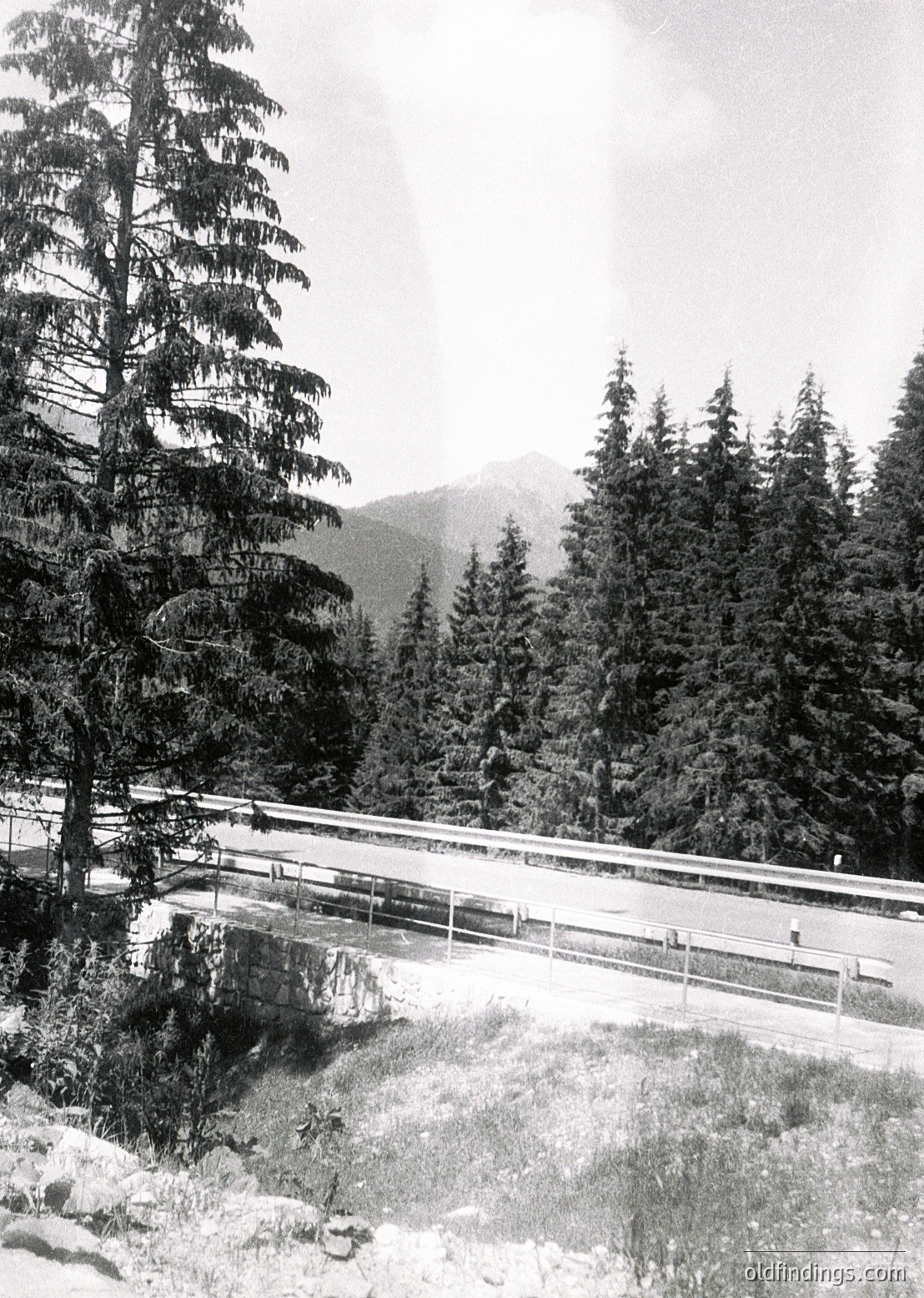 Snow-covered alpine road bridge spanning a rocky stream, flanked by dense coniferous forest. Distant mountain peaks and misty horizon suggest high-altitude terrain. Likely mid-20th century due to monochrome and composition style.