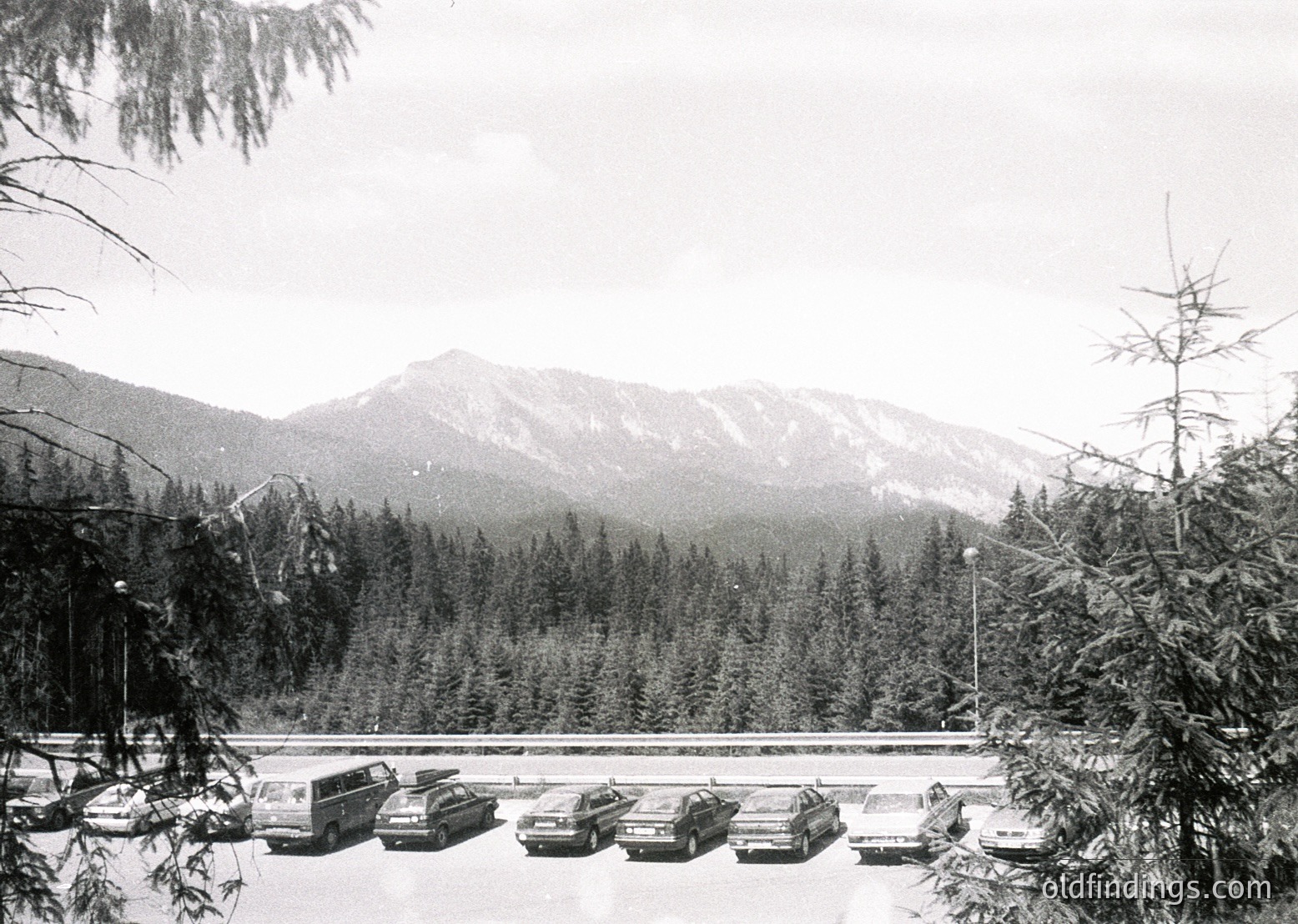 Mid-20th century mountain roadside parking area with classic cars, likely 1970s–1980s. Snow-covered pavement and evergreen forest framing rugged peaks.