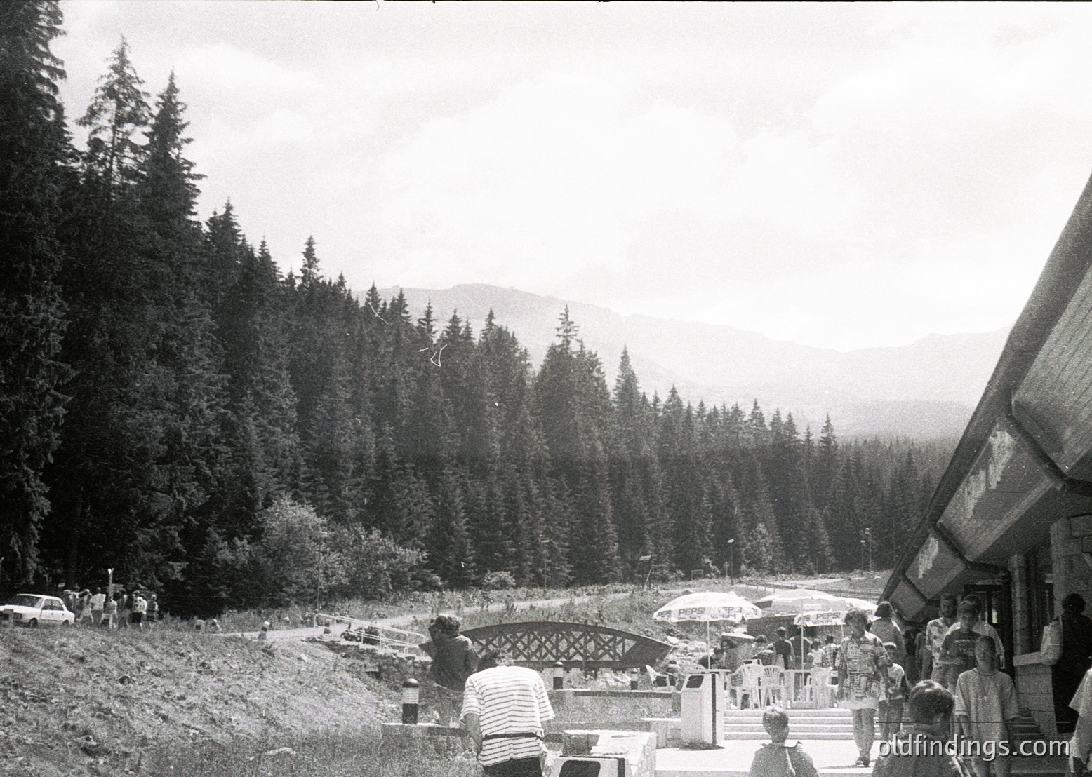 Mid-20th century alpine resort scene with dense coniferous forest and mountain backdrop. Wooden bridge and stone pathway lead to a lakeside area with wooden structures and outdoor seating. Crowded with mid-century attire (striped shirts, dresses, hats). Likely European alpine region.
