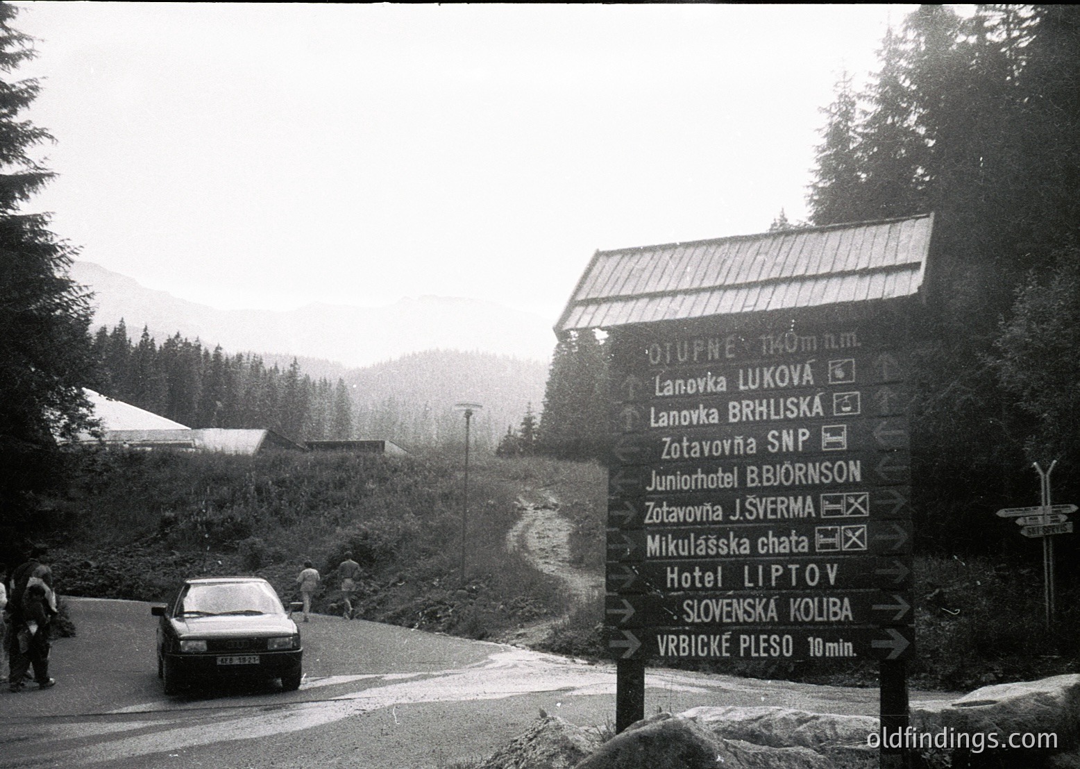 Black-and-white alpine road signpost listing destinations: Lanovka Lukova, Lanovka Brhliska, Zotavna J. Šverma, Mikuláška chata, Hotel Lípová, and Vrbické pleso (10 min). Sign indicates 1040m elevation. Snow-capped peaks and dense forest in background. Mid-20th century roadside scene, likely Slovakia or Czech Republic.